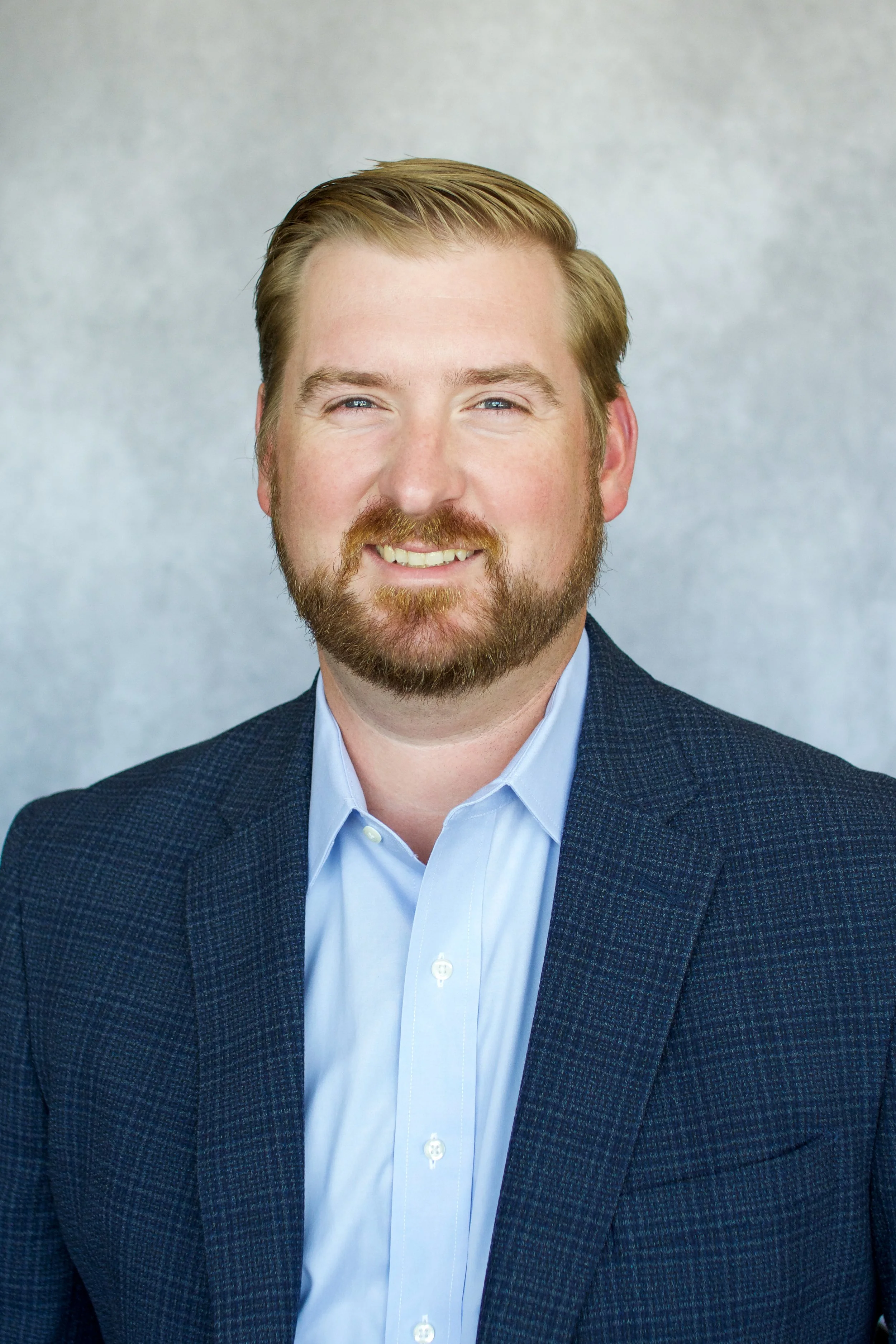Portrait of a man with light brown hair and a beard, wearing a dark blue blazer and a light blue shirt, smiling at the camera against a neutral background.