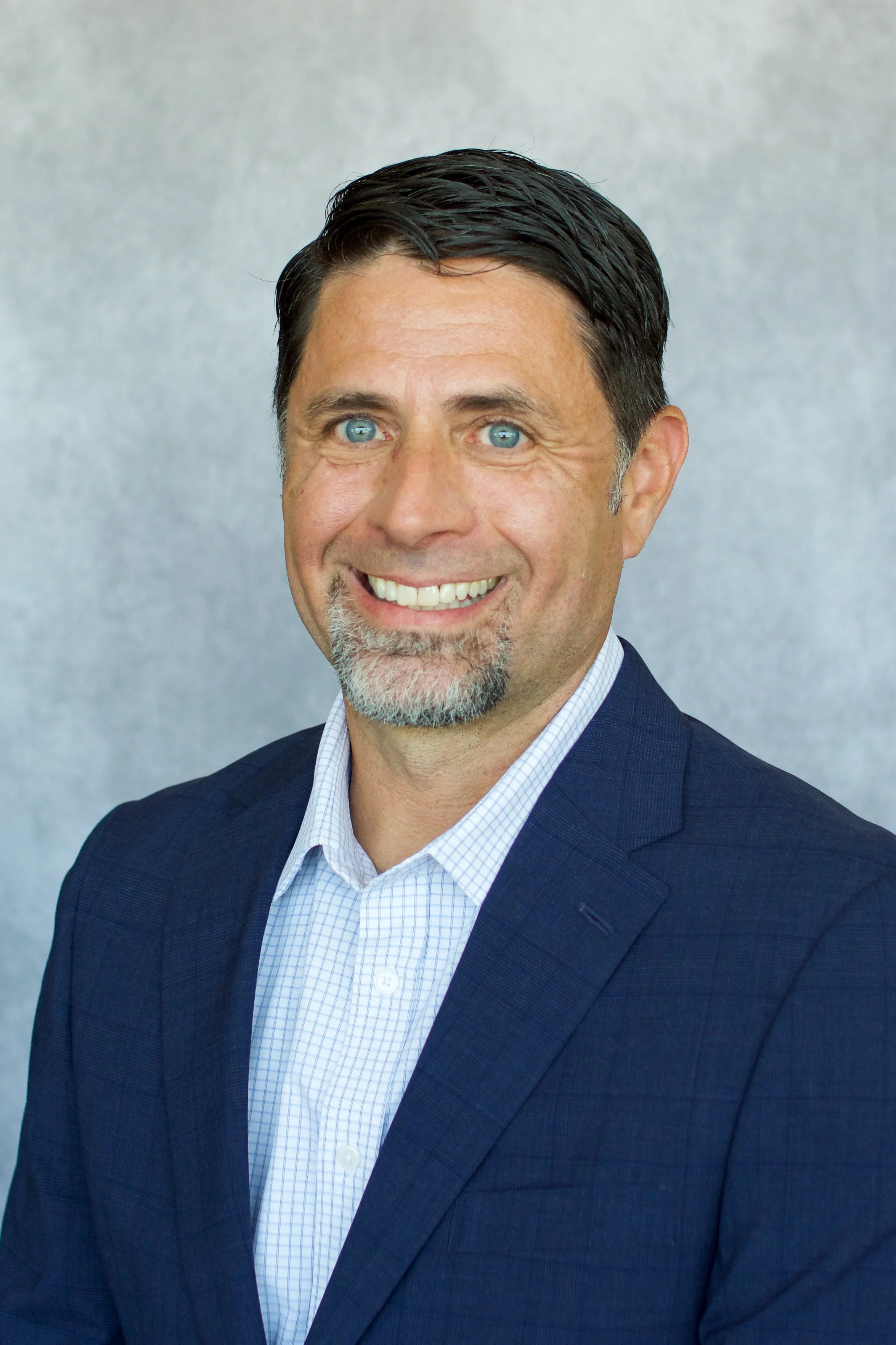 Professional headshot of a smiling man with dark hair, blue eyes, a beard, and a mustache, dressed in a navy blue suit and light blue checkered shirt, against a neutral gray background.