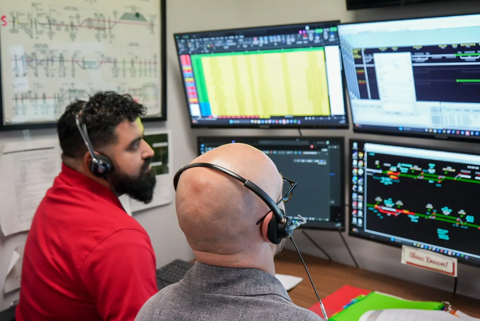 Two men, one with a beard and one bald wearing glasses, working on multiple computer monitors displaying financial data. They are in an office with charts on the wall.