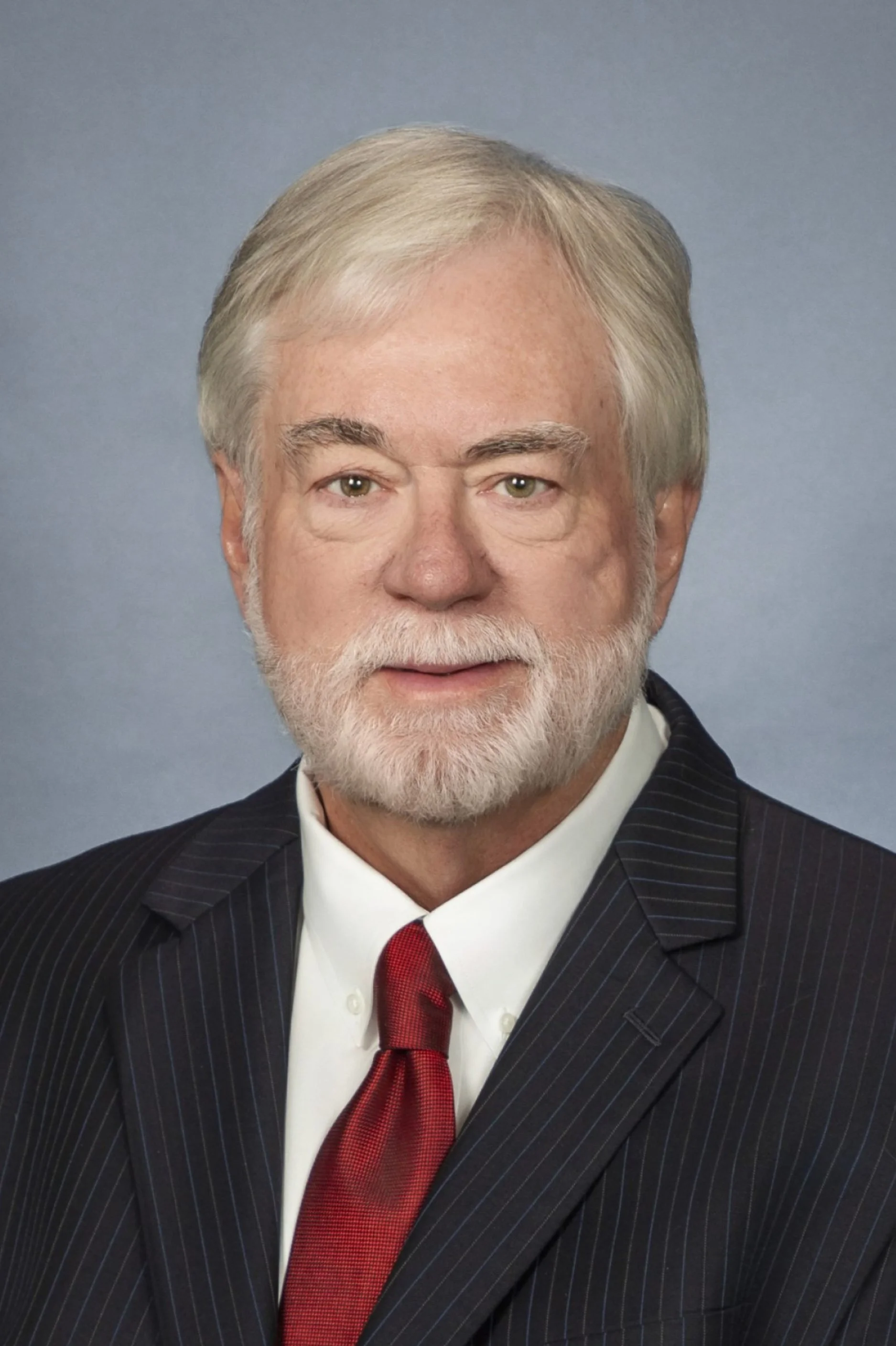 A professional headshot of an older man with white hair and a beard, wearing a dark pinstripe suit, white shirt, and red tie, against a light blue background.