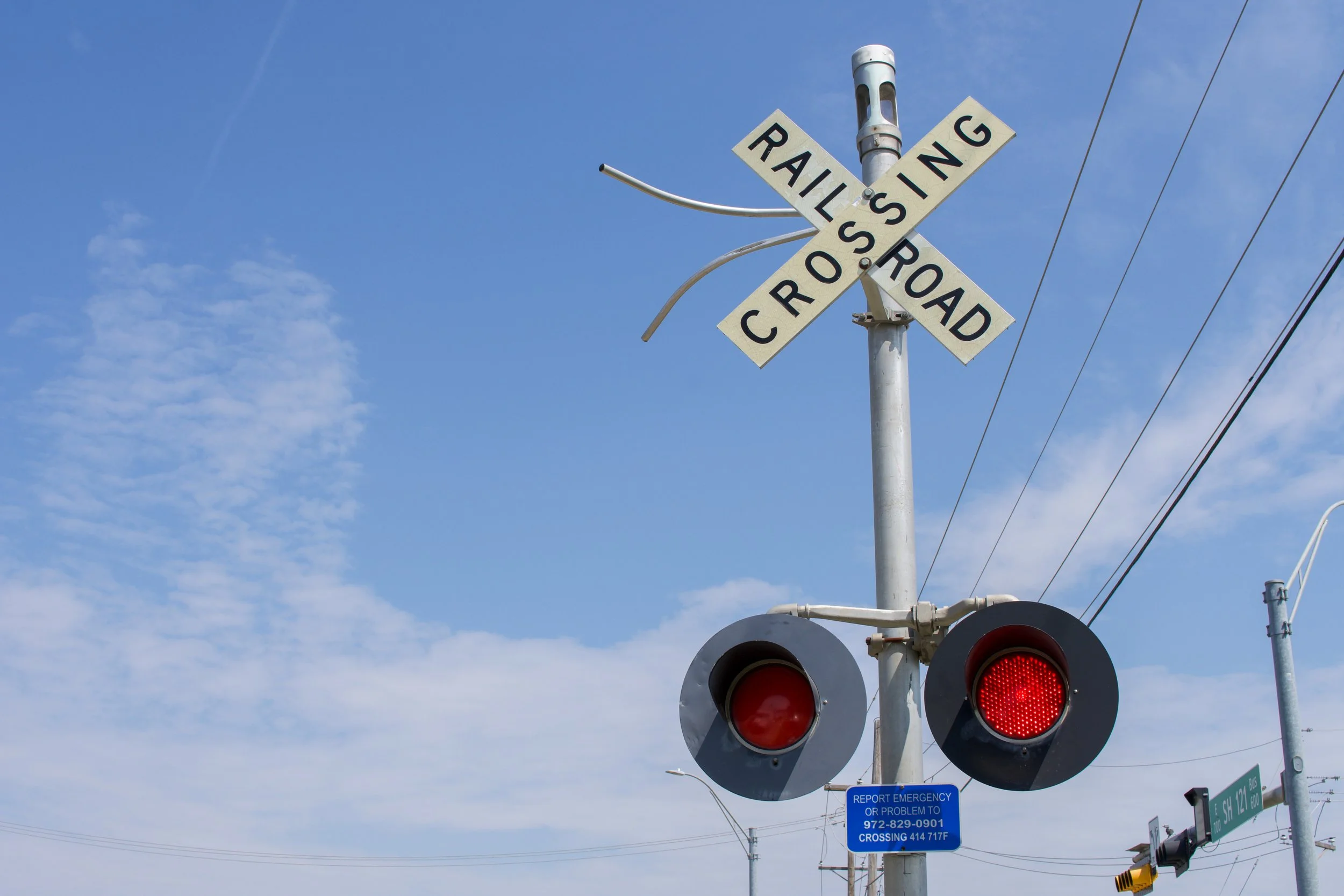 A railroad crossing signal with red lights flashing, a 'Railroad Crossing' sign, and an emergency contact blue sign mounted on a metal pole under a partly cloudy sky.