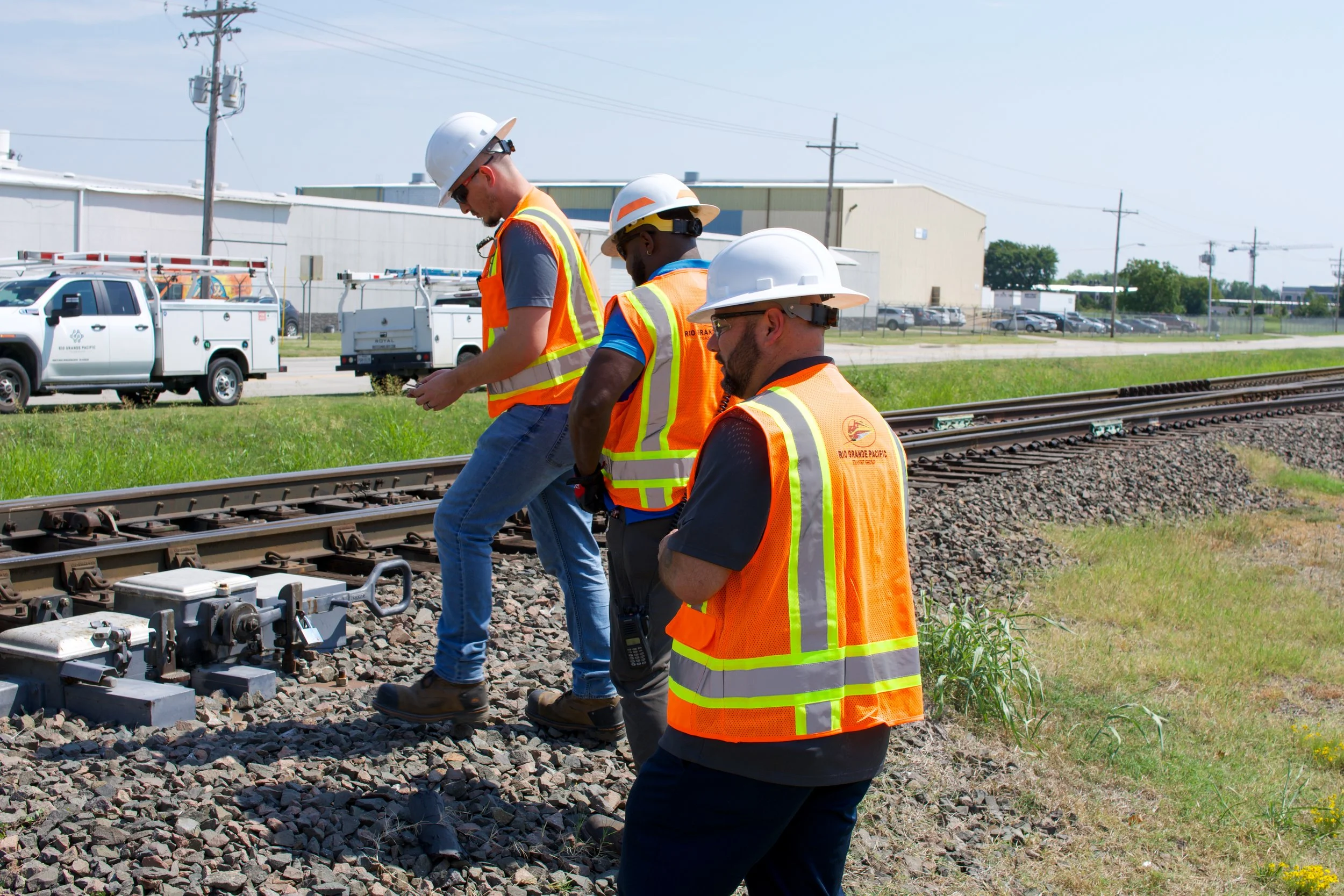 Four workers wearing safety helmets and high-visibility vests inspecting railway tracks on a sunny day.