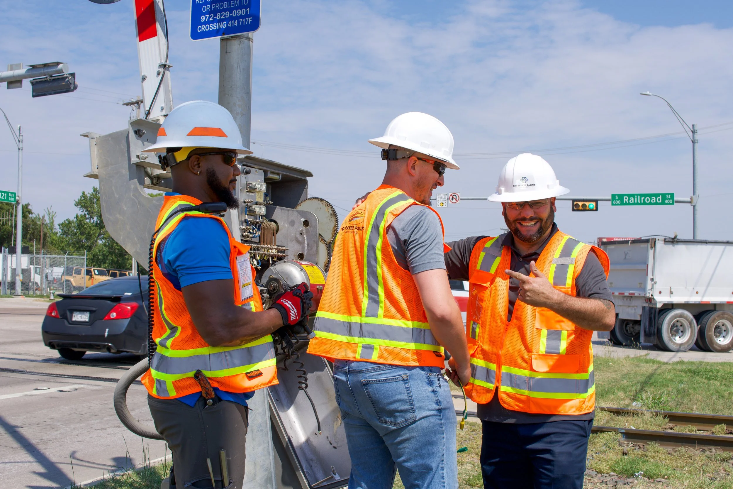 Three utility workers wearing safety helmets and orange reflective vests are standing near railroad tracks, talking and smiling under a clear sky with traffic signs and fencing in the background.