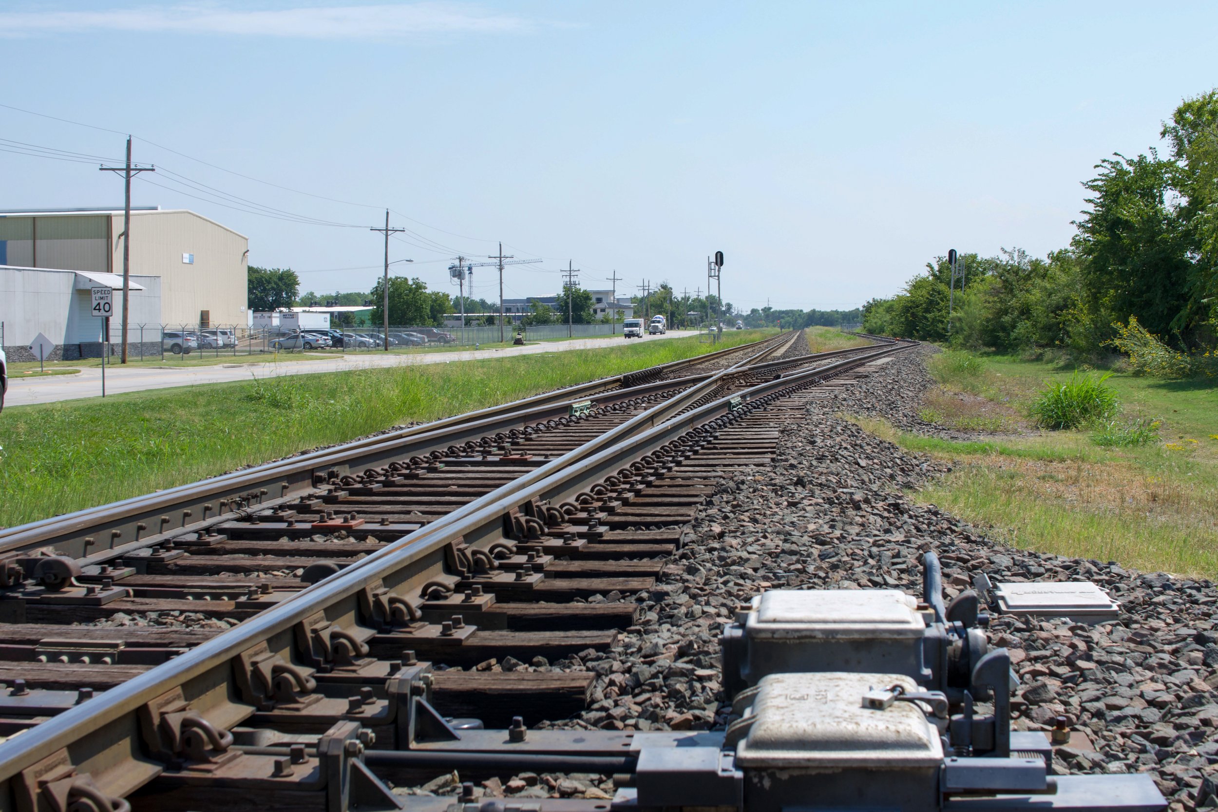 View of railway tracks stretching into the distance alongside a road with cars and industrial buildings on the left, and green trees and grass on the right, on a clear day.