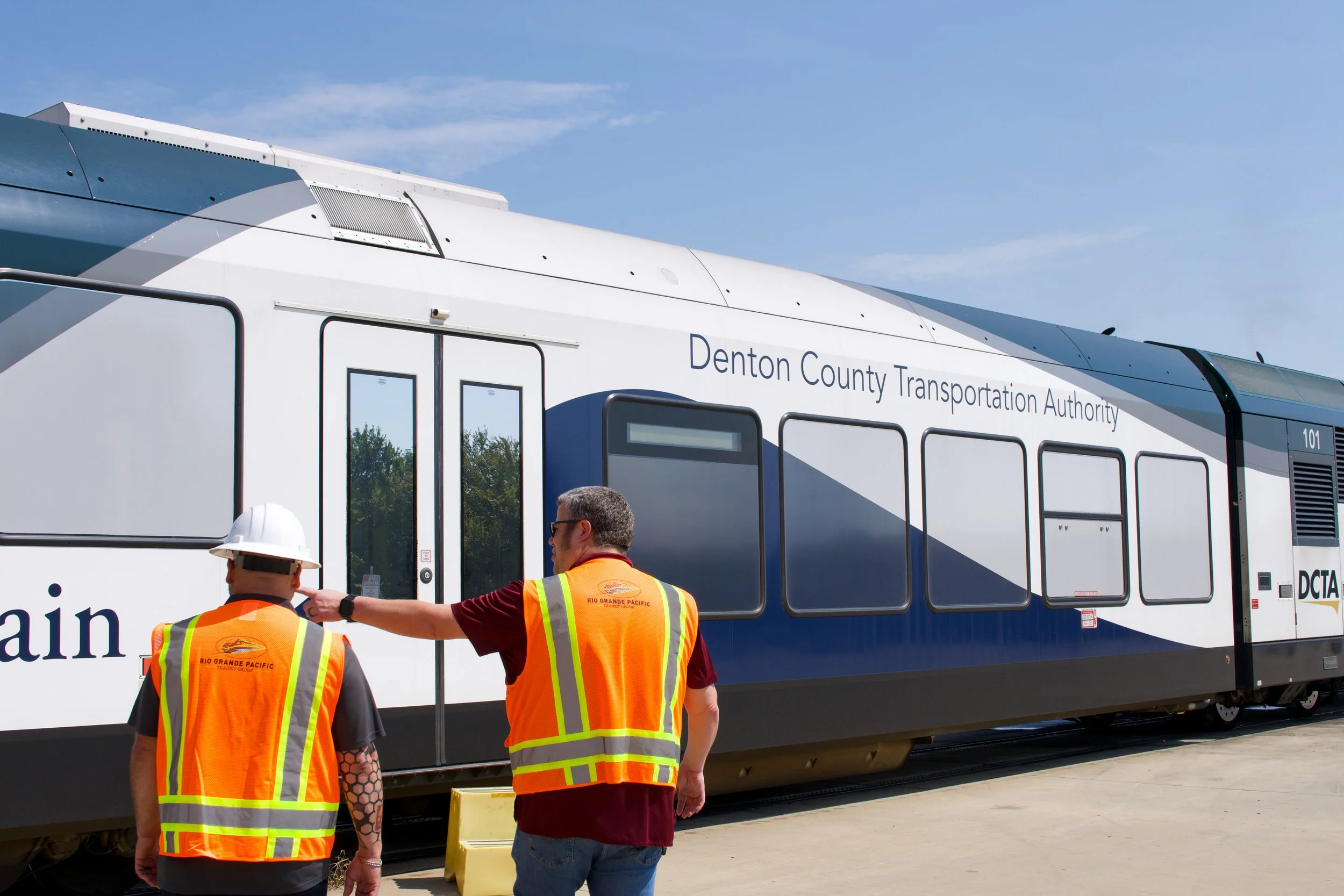 Two men in safety vests and one with a hard hat standing next to a modern train with the words "Denton County Transportation Authority" on it.