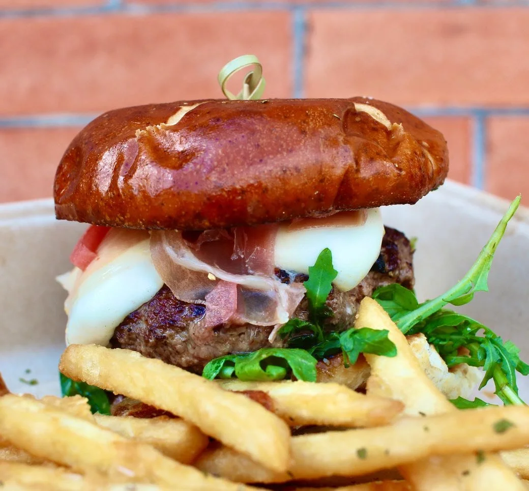Close-up of a gourmet cheeseburger with crispy fries on the side, featuring a toasted bun, melted cheese, a beef patty, ham, greens, and a toothpick.