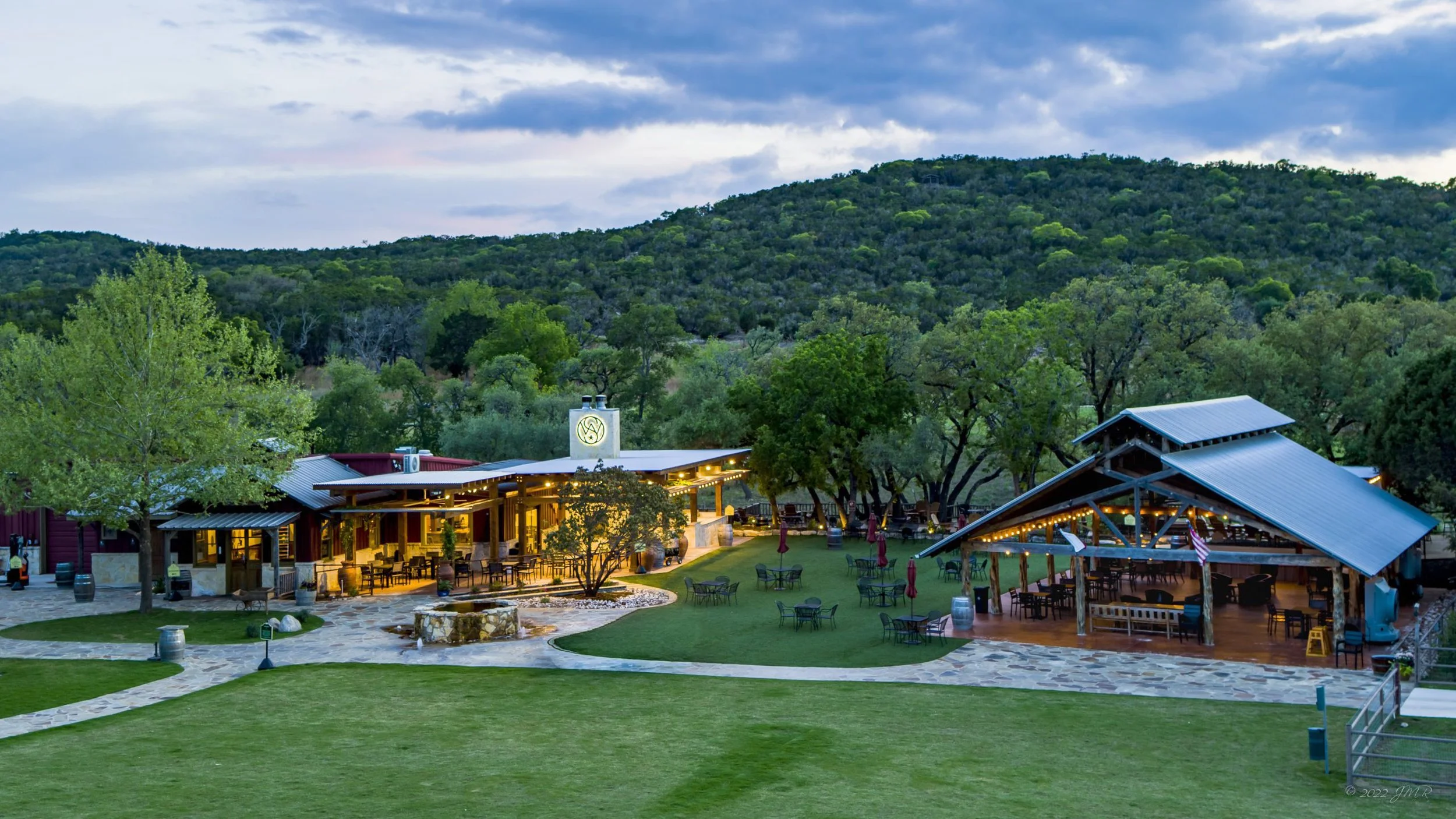 Outdoor restaurant or event space with string lights, tables, chairs, and a grassy area, surrounded by trees and hills, during dusk or evening.