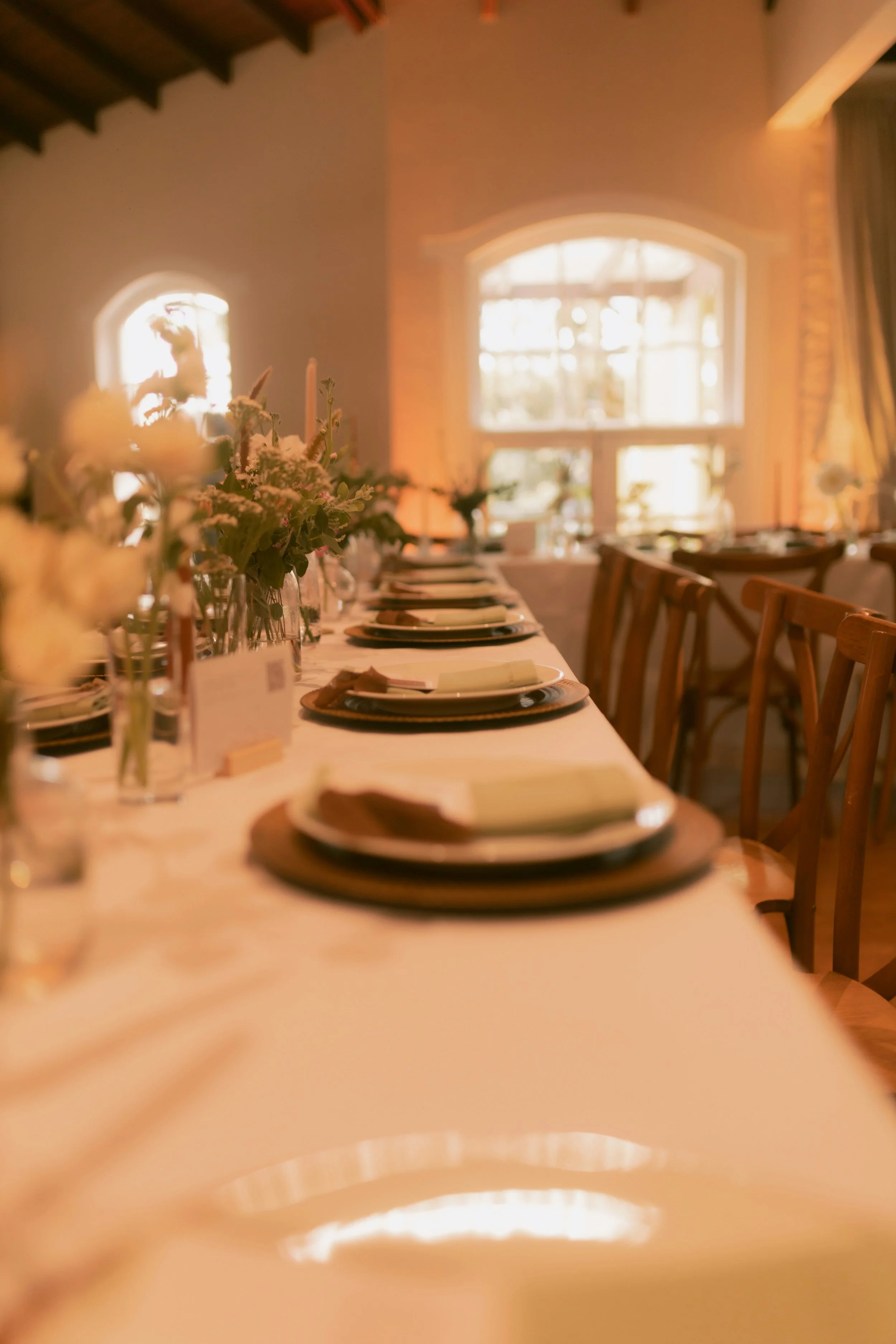 A long dining table set for a meal with plates, napkins, and glasses, decorated with floral centerpieces, in a warmly lit room with large windows and wooden chairs.