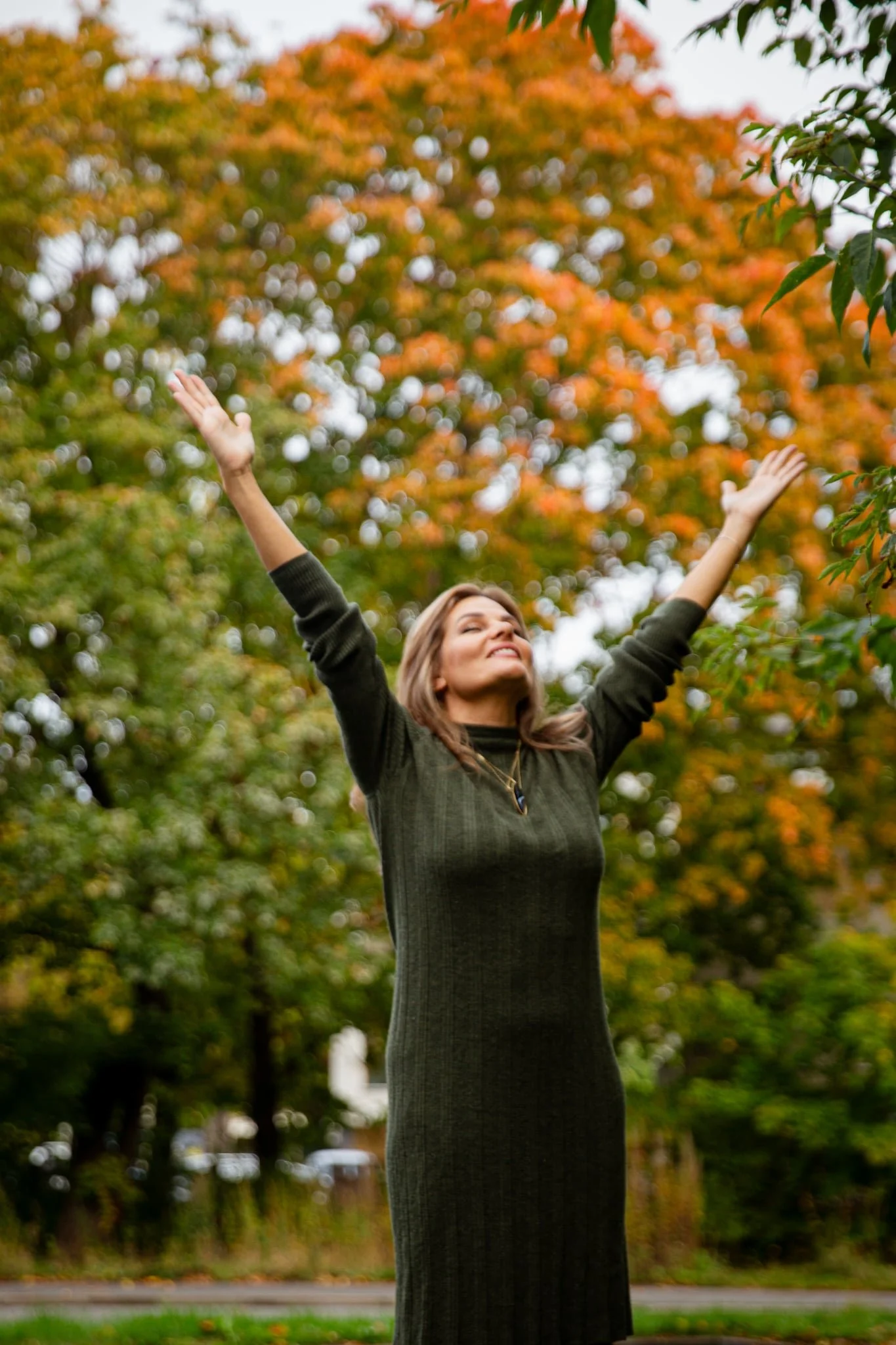A woman outdoors with arms raised, smiling, surrounded by autumn trees with orange, yellow, and green leaves.