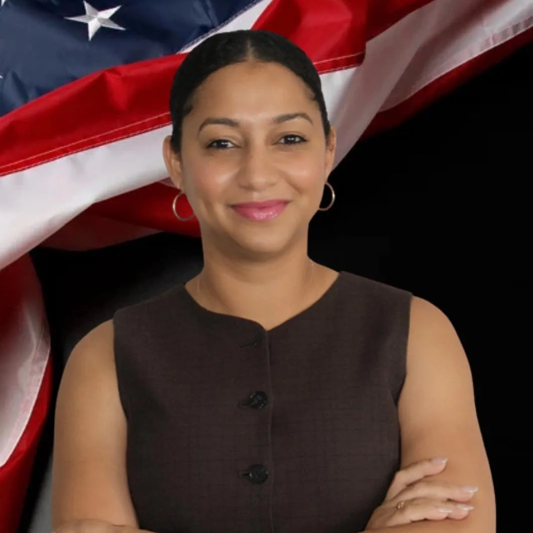 A woman with short dark hair and hoop earrings, wearing a sleeveless dark top, standing in front of an American flag backdrop.