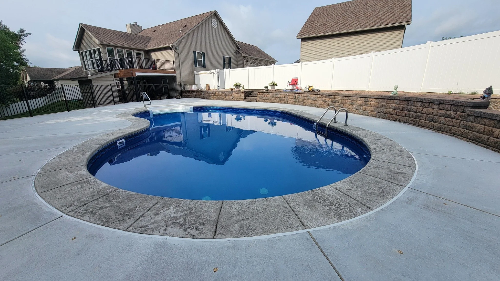 Backyard with a small in-ground swimming pool, concrete deck, and a white privacy fence. Houses with decks and a brick retaining wall are visible in the background.
