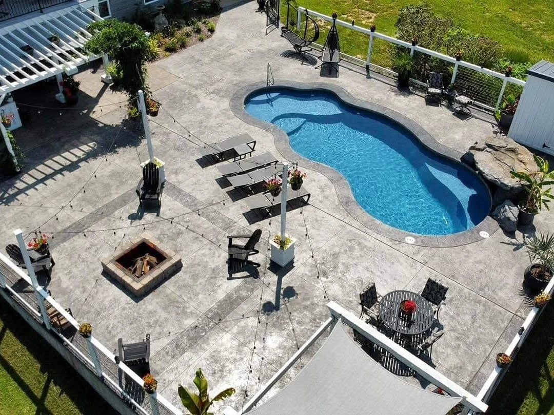 A backyard patio with a small kidney-shaped swimming pool, surrounded by outdoor furniture including benches, chairs, and a round table with chairs. There is a fire pit in the center and potted plants around the patio, with a fence and grassy lawn in
