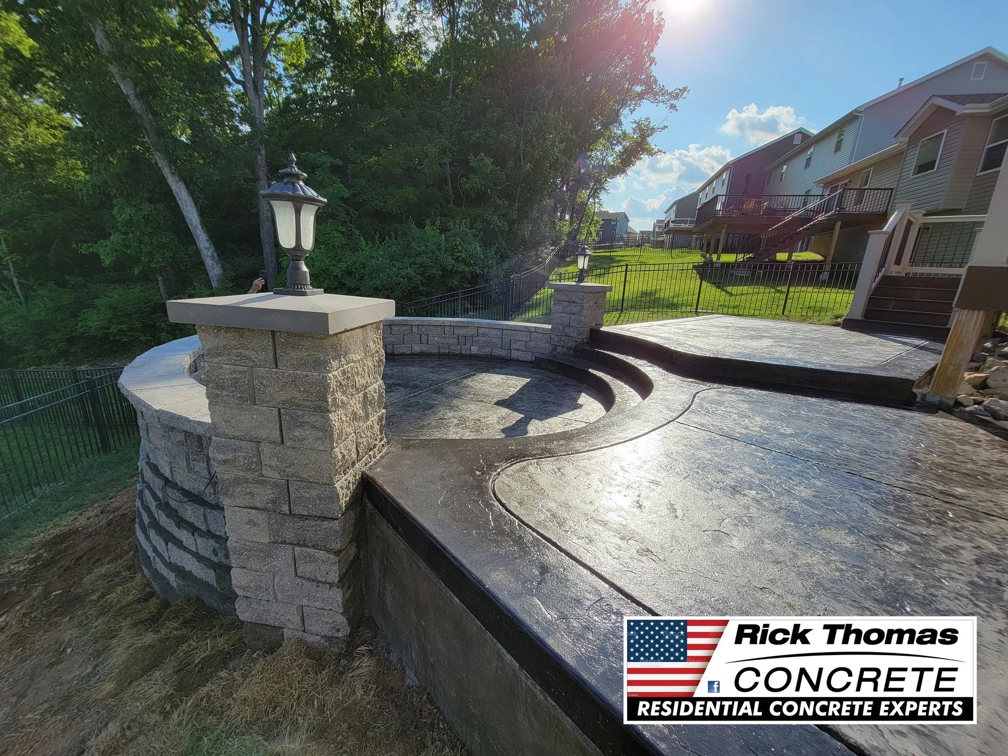 Newly poured concrete patio with a decorative stone wall and lampposts, overlooking a grassy backyard with houses in the background.