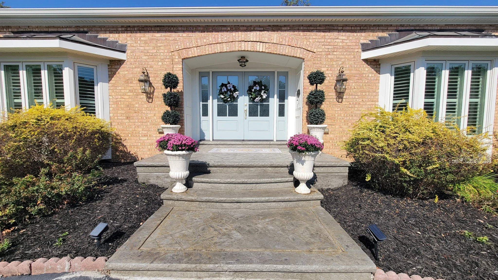 Front porch of a brick house with white double door decorated with two wreaths. Two white planters with pink flowers on steps leading to porch. Bushes on either side of porch, solar lights on ground.