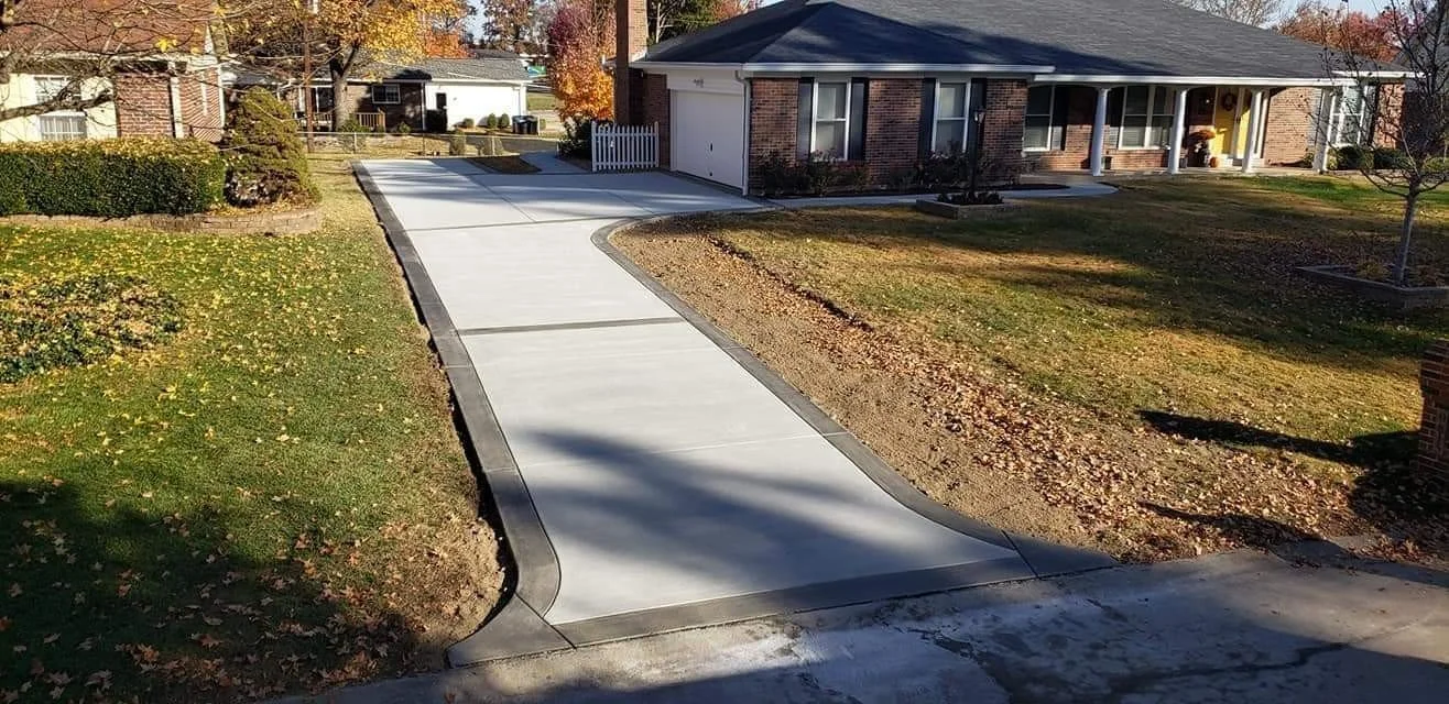 New concrete driveway and sidewalk leading to a brick house with a covered porch, in a suburban neighborhood with trees and lawns, during autumn.