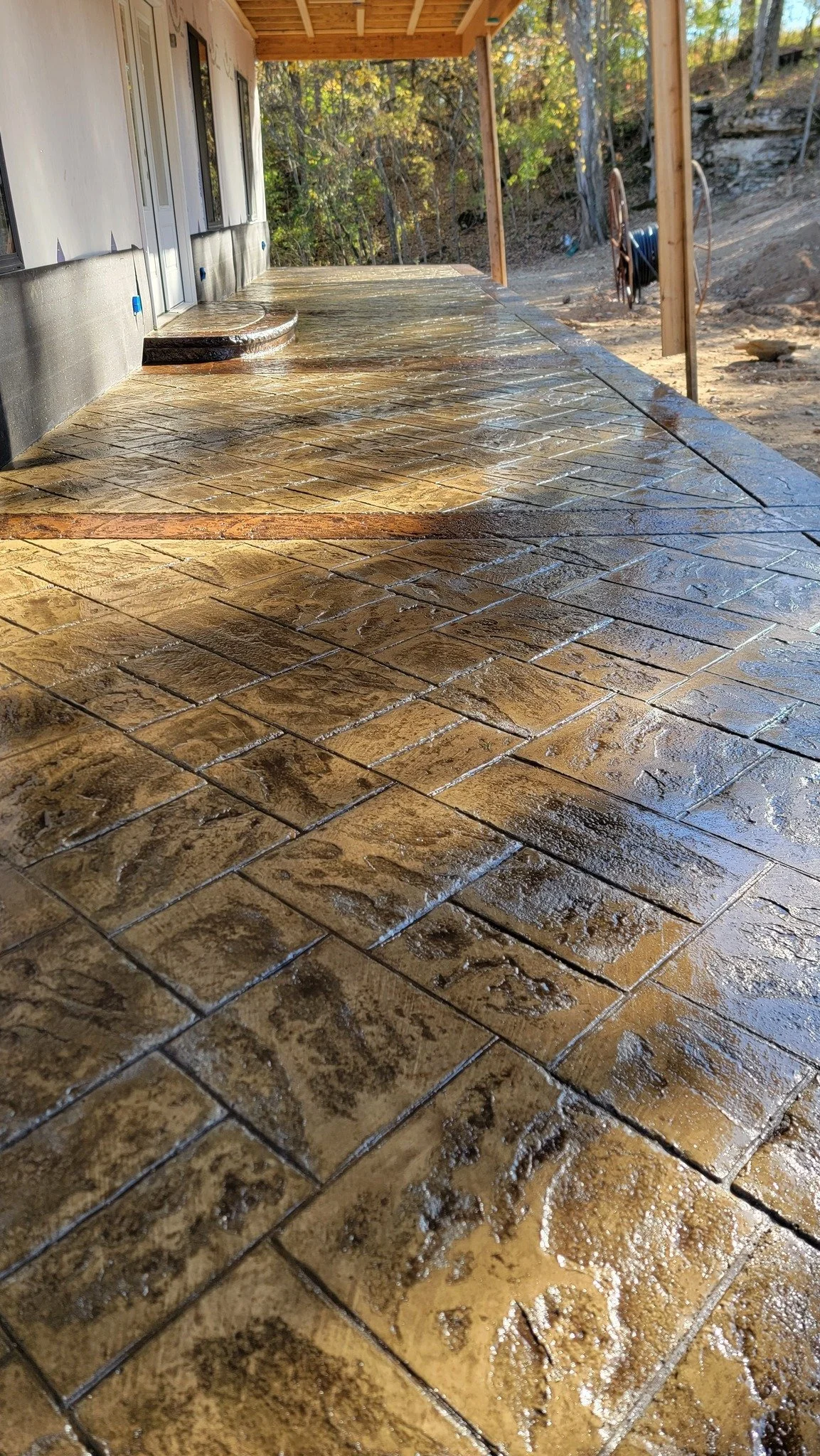 Close-up of a freshly sealed concrete patio with wood-like stamped design, attached to a house with sliding glass doors, overlooking a wooded backyard.