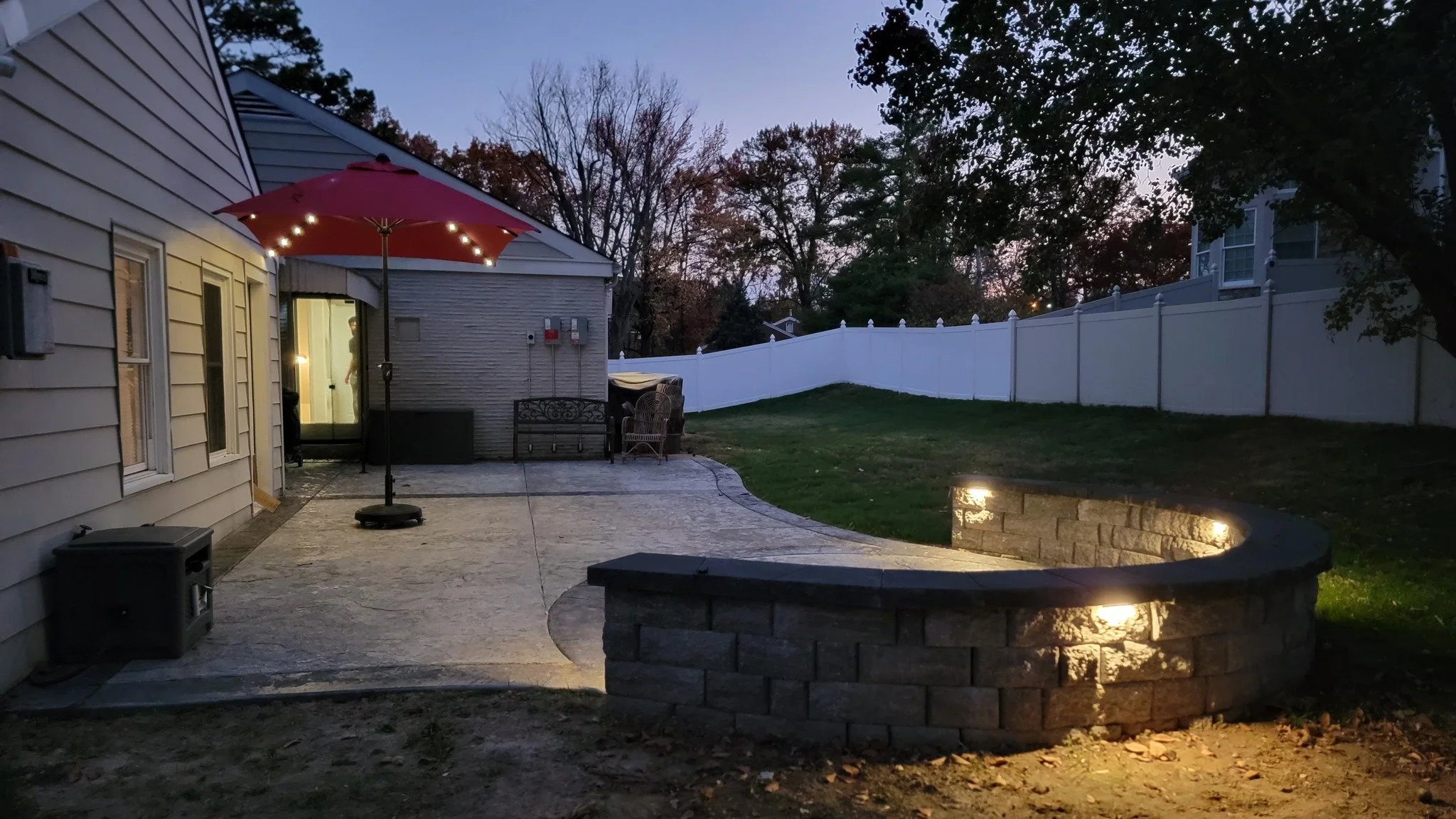 Backyard patio area at dusk with a red umbrella, a lit fire pit, a white privacy fence, and trees in the background.