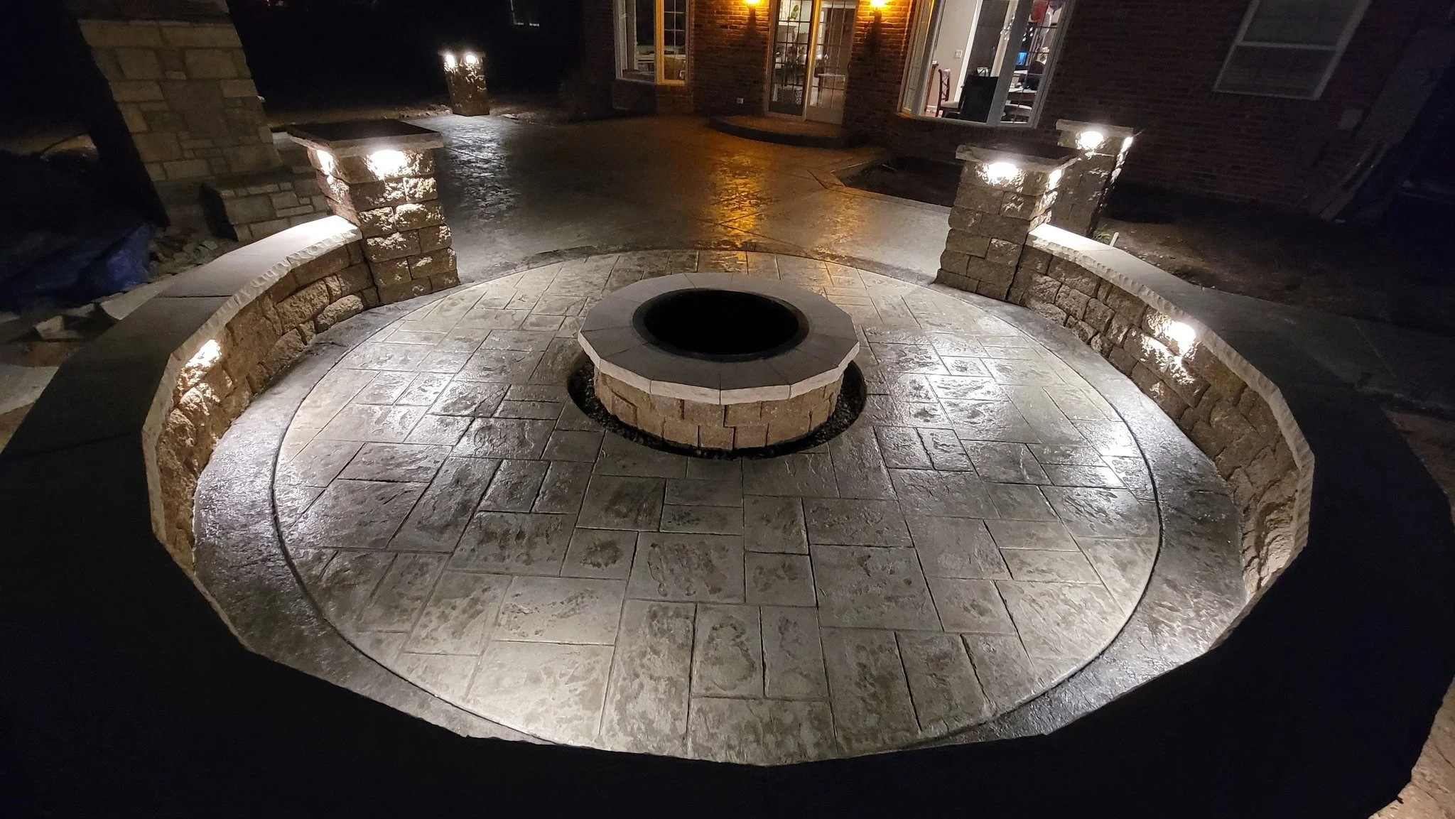 Nighttime view of a circular paved area with a raised fire pit in the center, surrounded by stone benches built into brick pillars, with lights illuminating the area.