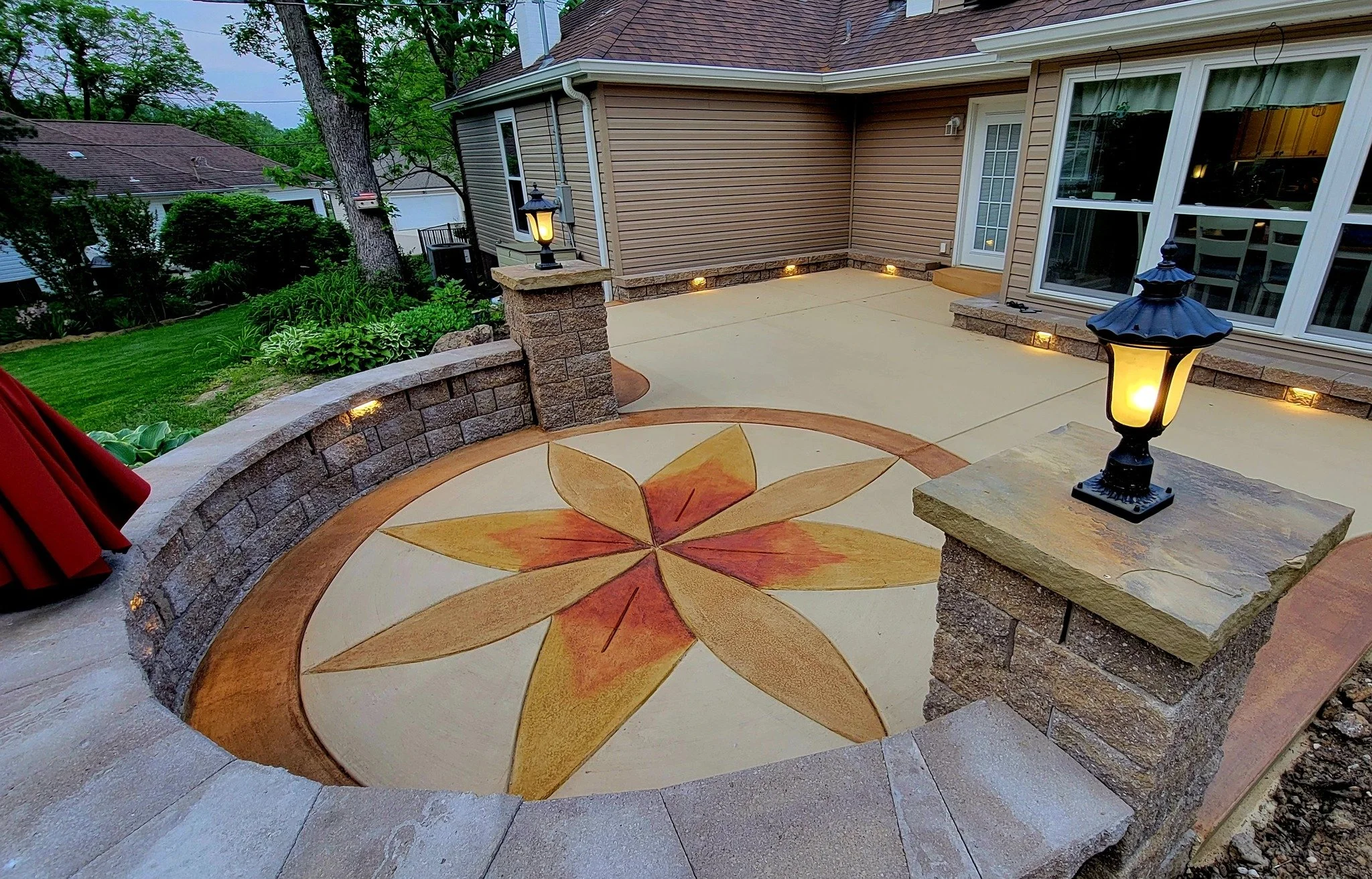 Backyard patio featuring a decorative circular design with a star pattern in the center, surrounded by stone retaining walls and illuminated by outdoor lanterns at dusk.
