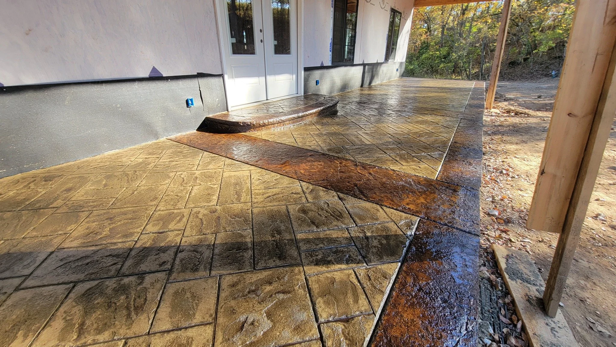 Newly installed concrete patio with stamped and stained patterns, next to a house under construction, with trees in the background.