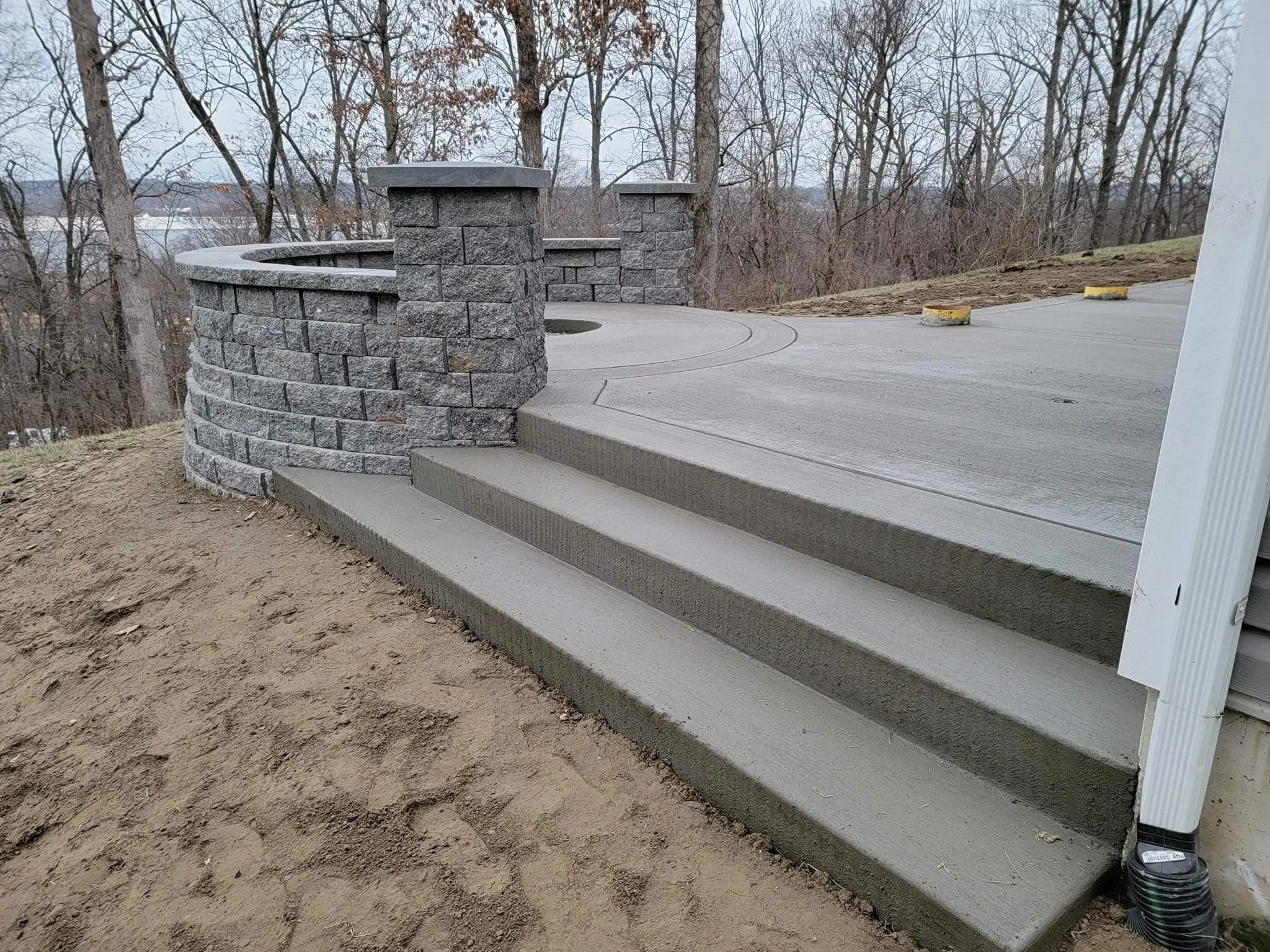Concrete steps leading to a paved driveway with a decorative stone wall on the left side, in a wooded outdoor area.