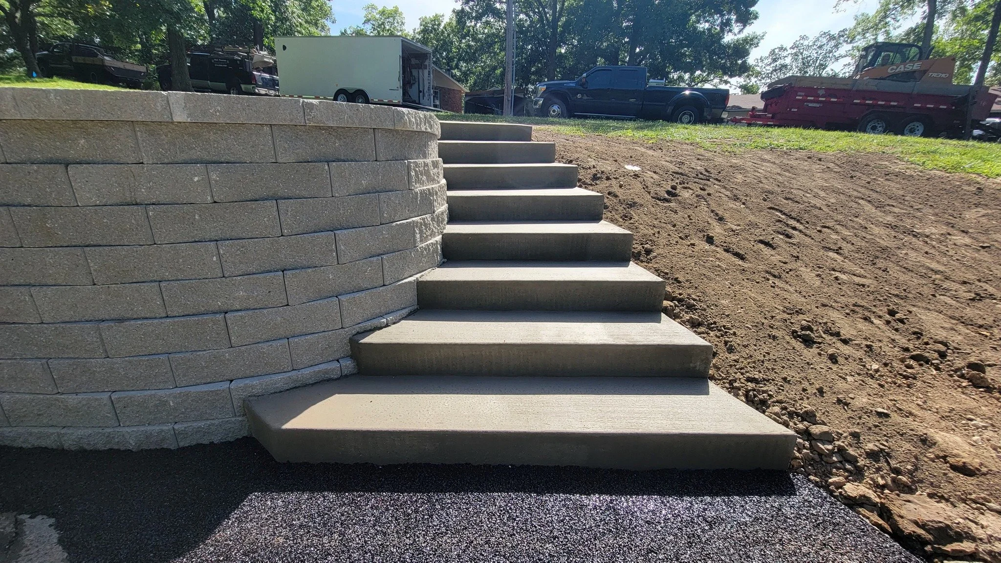 Concrete steps leading up to a raised brick structure at an outdoor construction site with trucks and trees in the background.