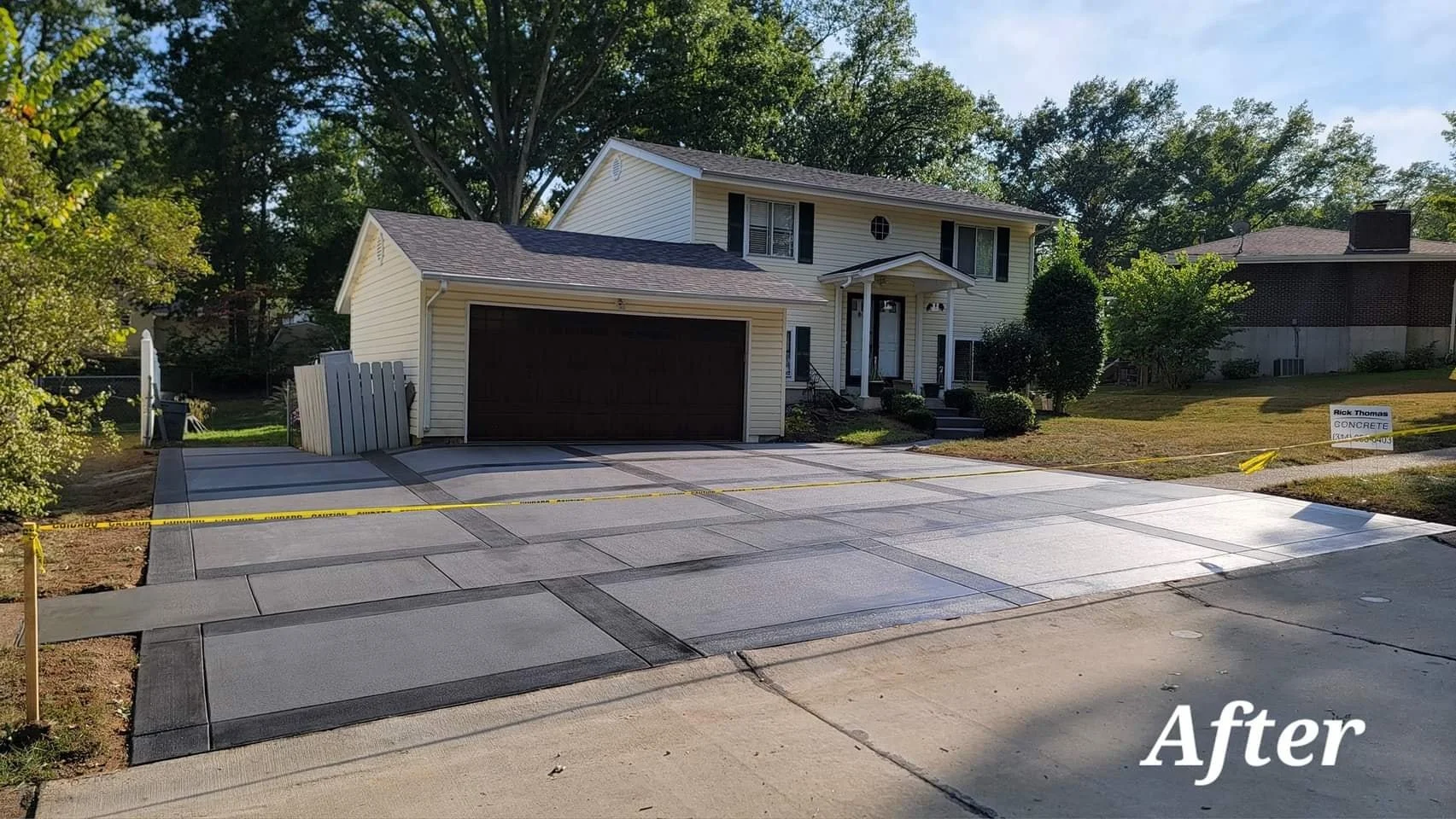 Newly installed decorative concrete driveway in front of a two-story house with a garage. The house has beige siding, black shutters, and a small front porch. There is a yard with some bushes and trees, and a "Concrete" sign on the lawn. The word "Af