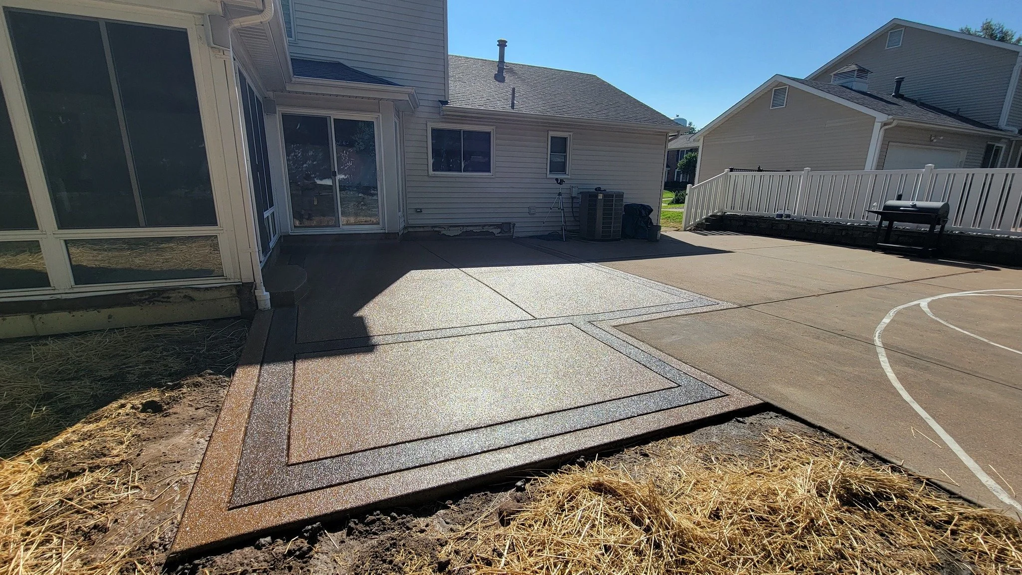 Backyard patio with concrete and textured flooring, a white house with sliding glass door and windows, a white fence, and a barbecue grill, on a sunny day.