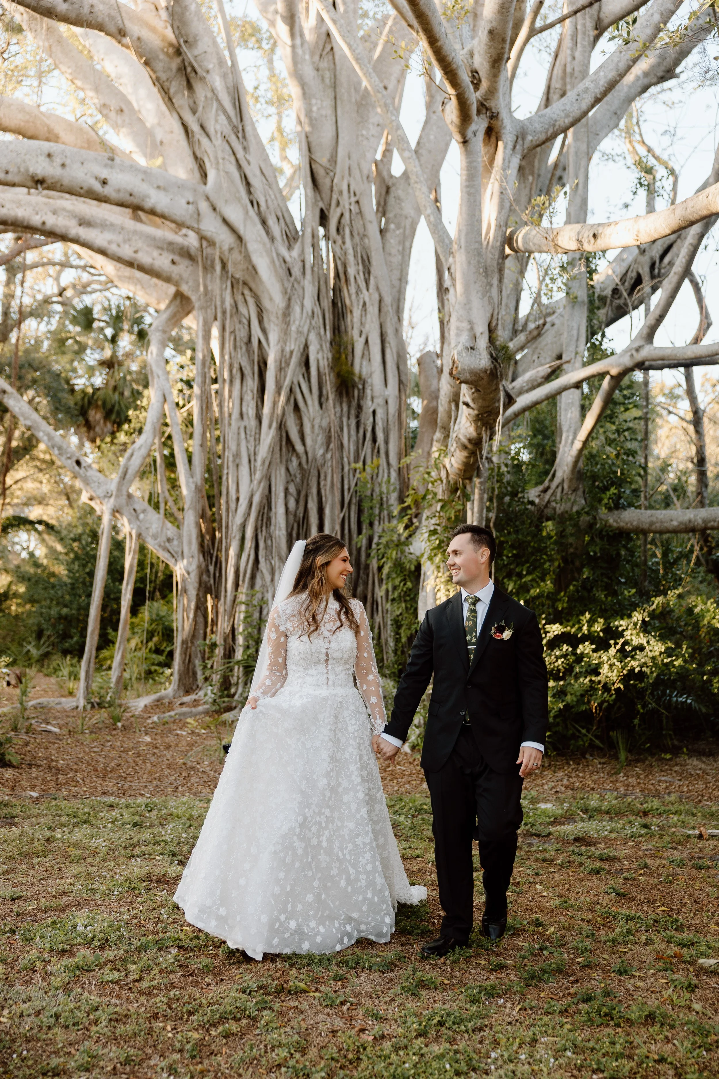 A man and woman holding hands in a grassy field, with the man wearing a suit and cowboy hat and the woman in a white dress, during sunset.