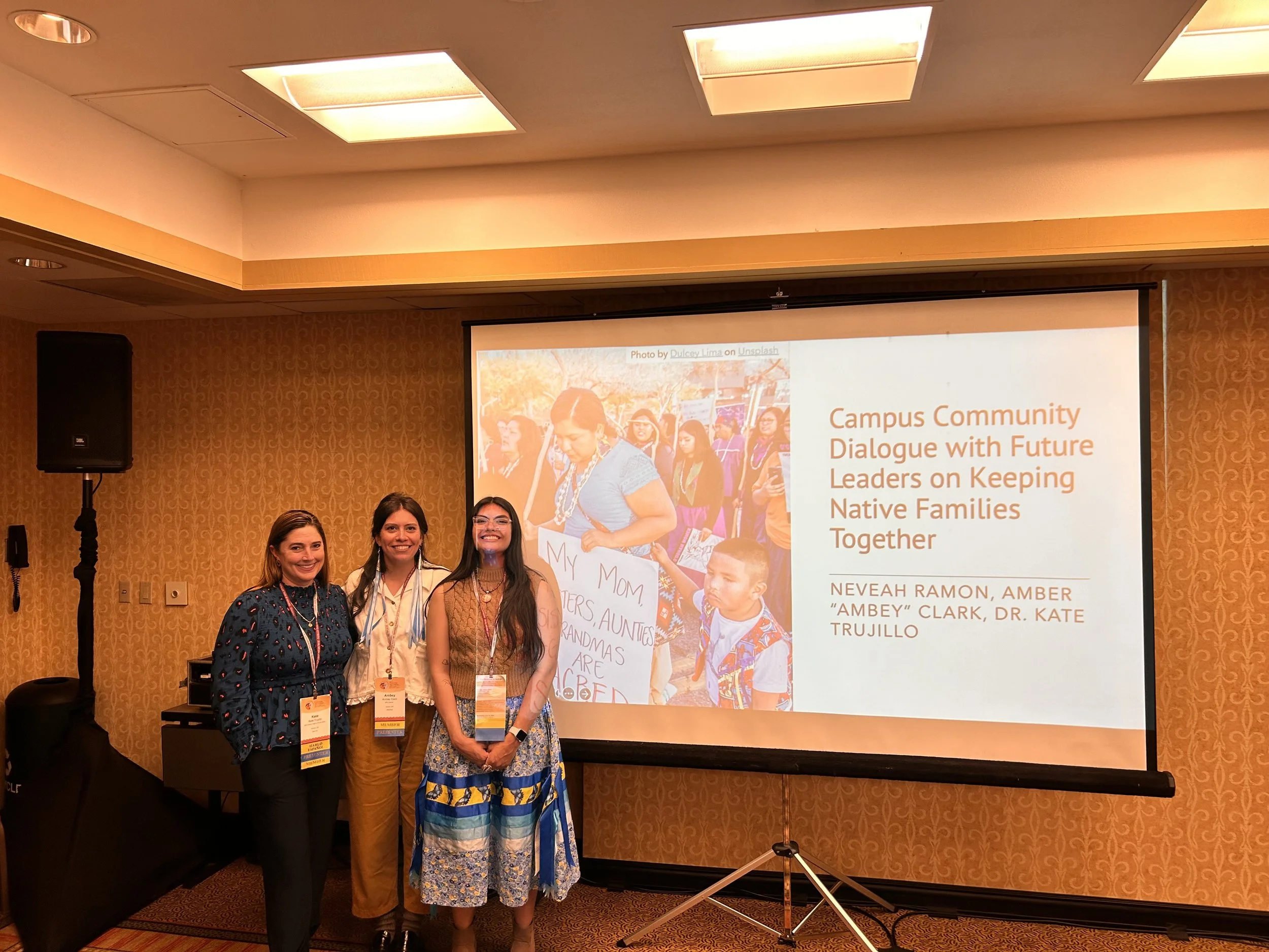 Nevaeh Ramon (far right) stands beside two women (Dr. Kate Trujilo - left, and Ambey Clark - center). They are standing in front of a projected screen that reads "Campus Community Dialogue with Future Leaders on Keeping Native Families Together"