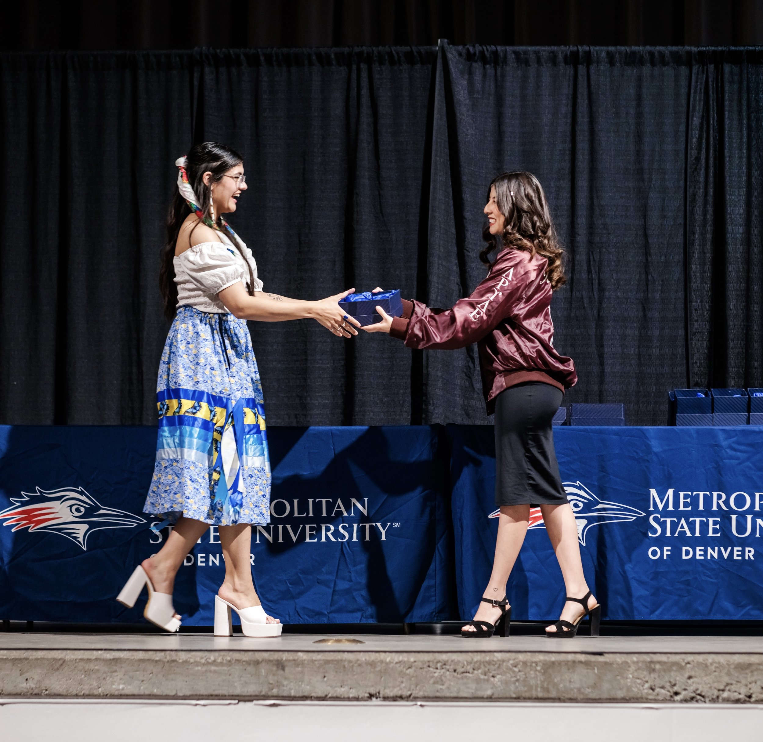 Nevaeh Ramon walking across a stage, reaching to receive an award handed to her by a second woman. Behind them is a branded banner with "Metropolitan State University of Denver" printed on.