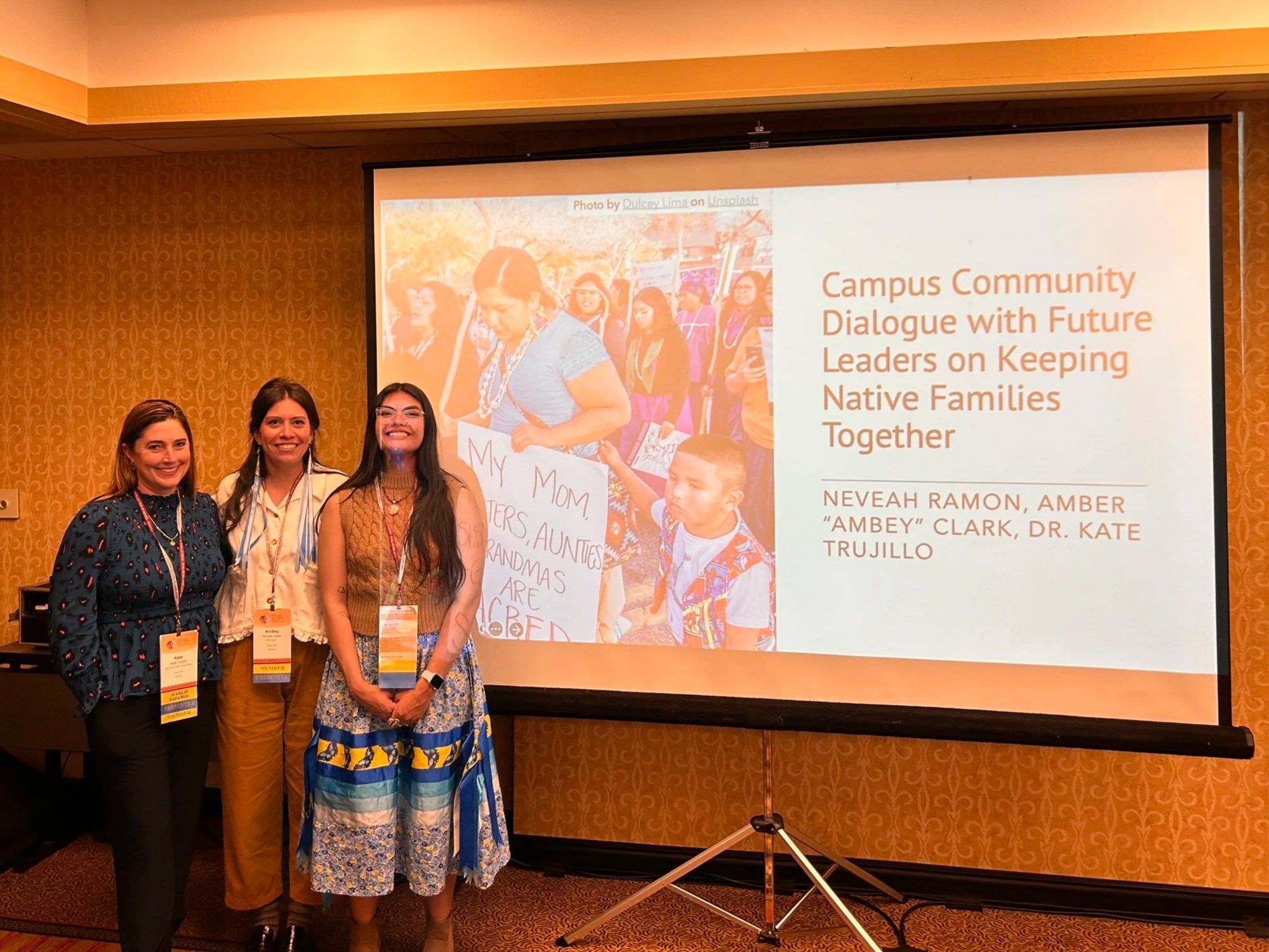 Three women stand in a row to the left of a projected screen reading "Campus Community Dialogue with Future Leaders on Keeping Native Families Together --- Nevaeh Ramon (Far right), Amber "Ambey" Clark (middle), Dr. Kate Trujilo (far left)