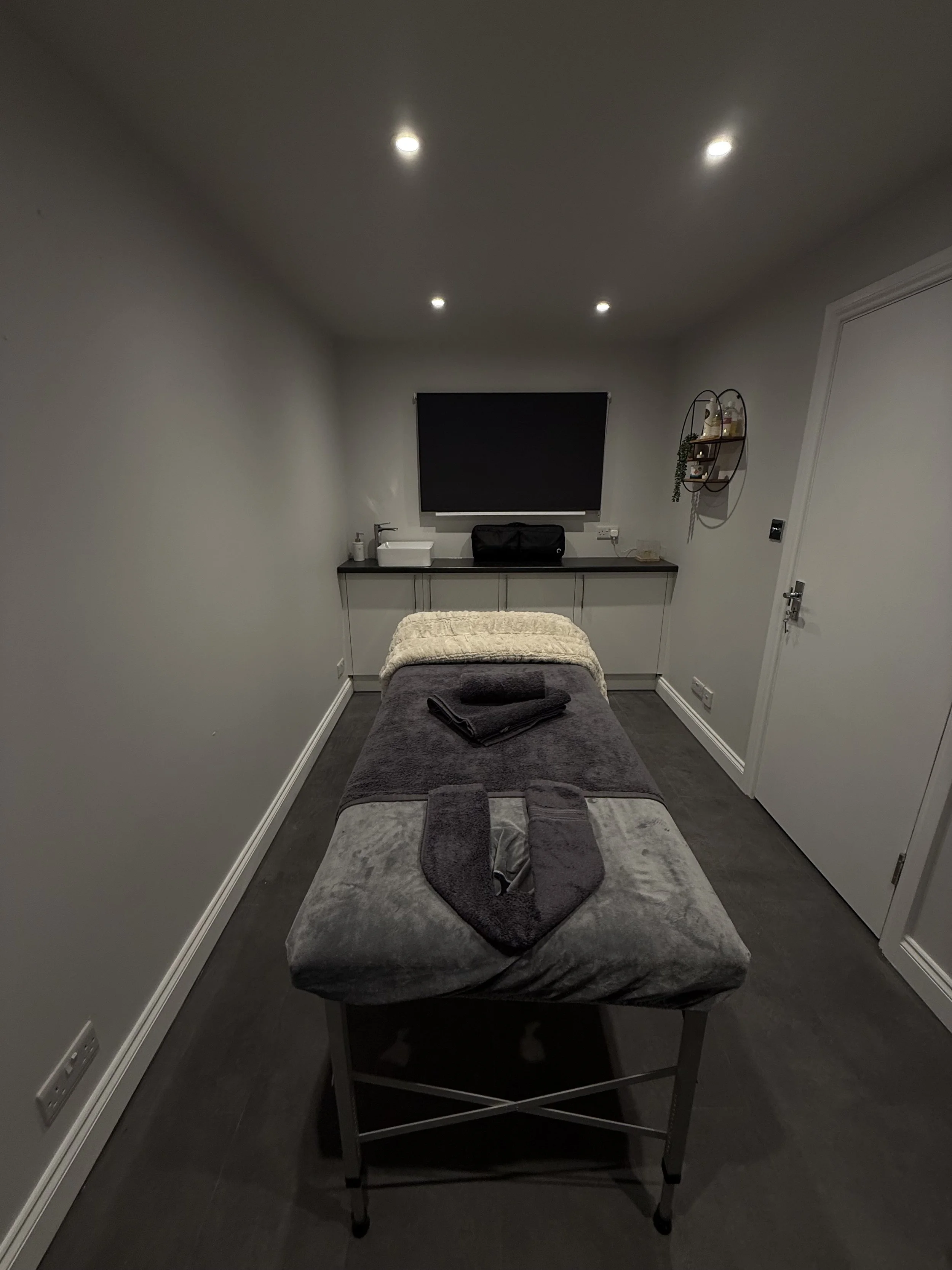 A massage room with a massage table covered in gray and beige towels, with folded black towels on top, in front of a cabinet with a sink and a black box, with a window and a decorative wall shelf holding small plants and bottles.