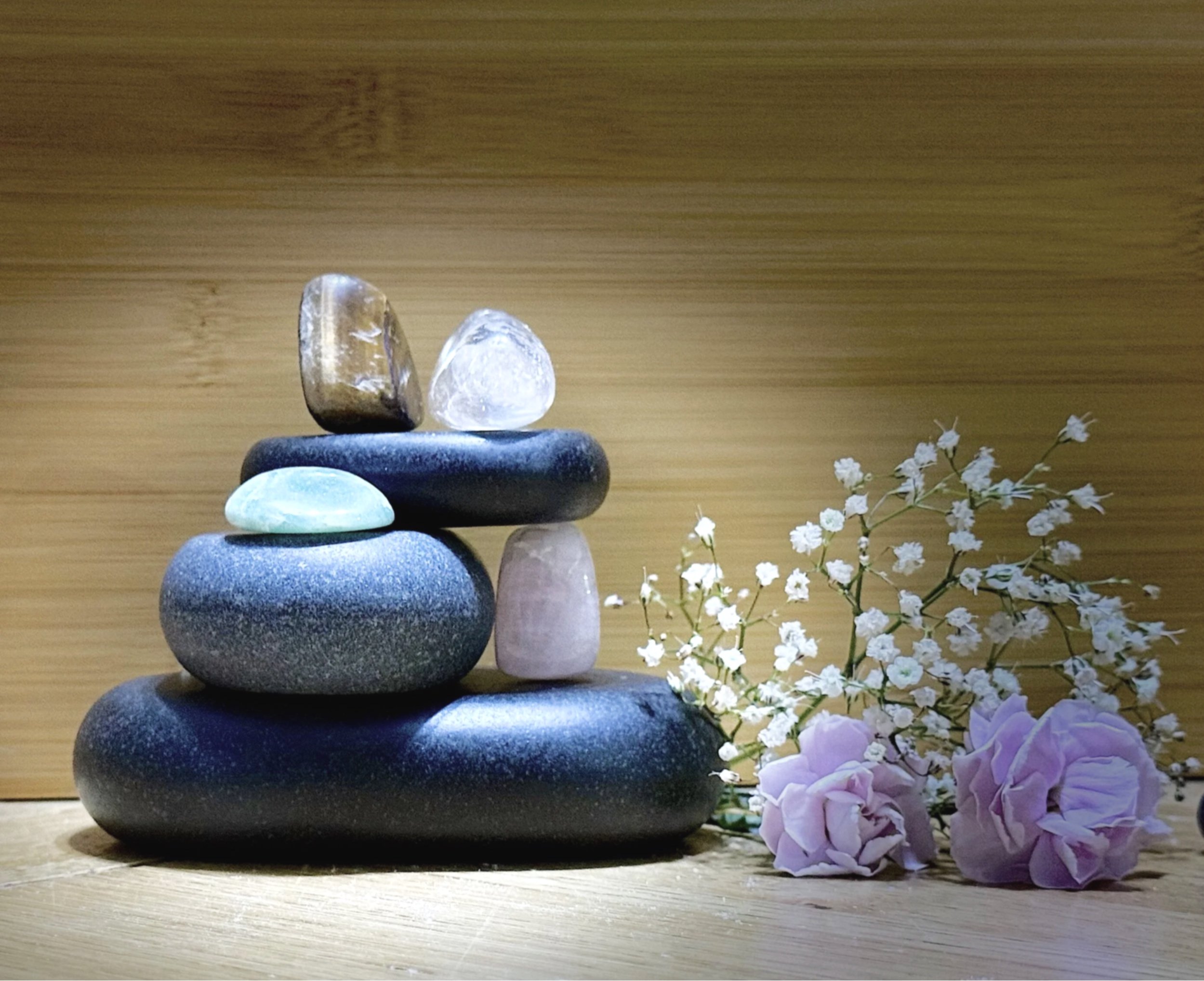 Stacked black stones with glass and mineral objects on top, flowers on the right, against a wooden background.