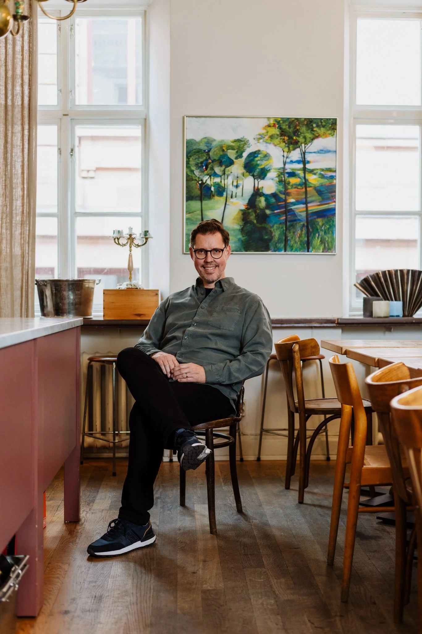 Man står i ett kök, klädd i grå skjorta och svarta byxor, framför ett bord och stolar, med ett färgglatt konstverk på väggen bakom. Ljusa fönster släpper in mycket ljus.