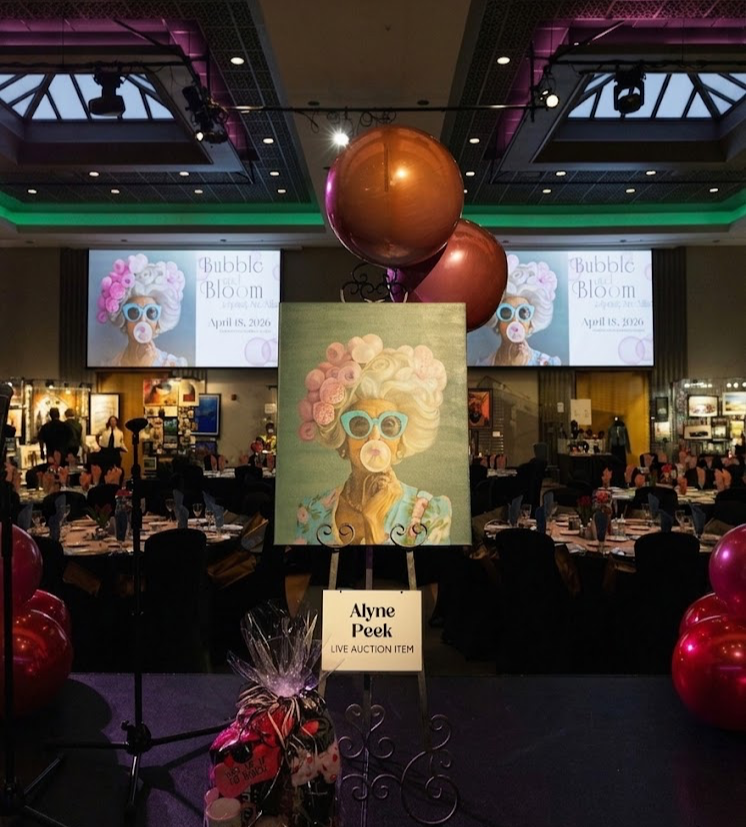 Auction event with a colorful painting of a woman with curly hair and sunglasses, balloons, and a large screen displaying similar artwork and event details.