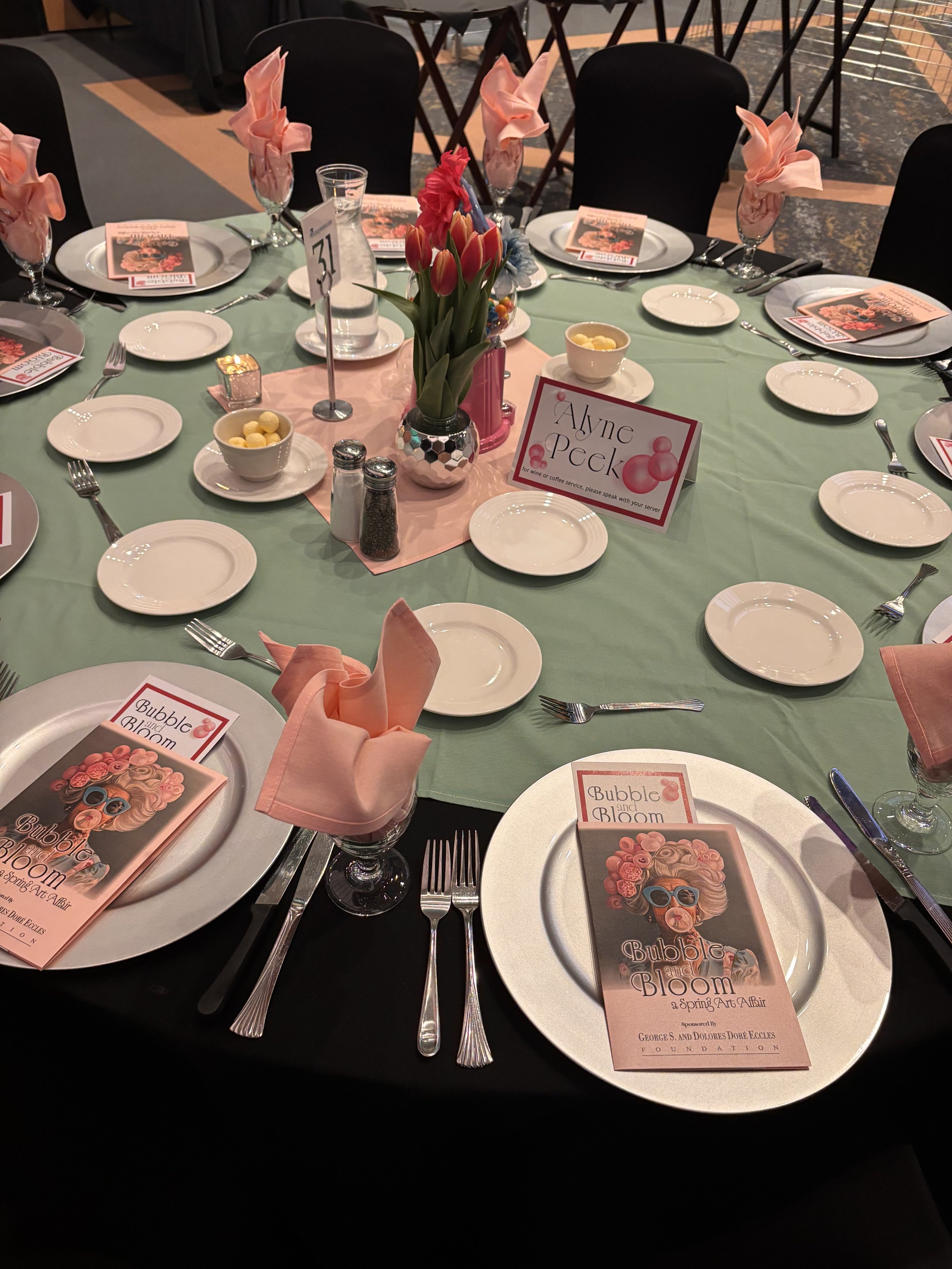 Round banquet table set for a formal event with black chairs, pink napkins folded in glasses, white plates, silverware, small bowls of candy, a vase of pink and red flowers in the center, event programs titled 'Bubble and Bloom,' and a table card with the name 'Ayne Peck.'