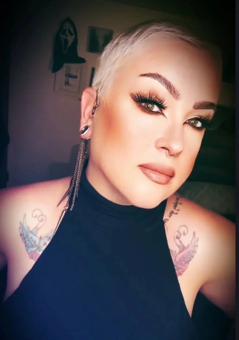 A woman with short platinum blonde hair, dramatic makeup with dark eyeliner and long lashes, wearing a black sleeveless top, earrings with chains, and showing tattoos on her shoulders.