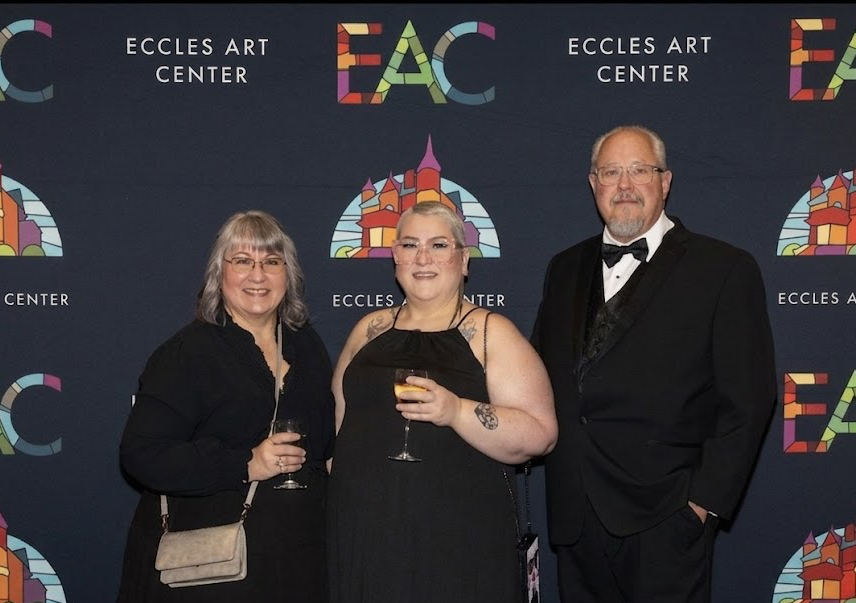 Three people smiling and holding wine glasses at an art event, standing in front of a backdrop with 'ECCLES ART CENTER' and colorful logo.