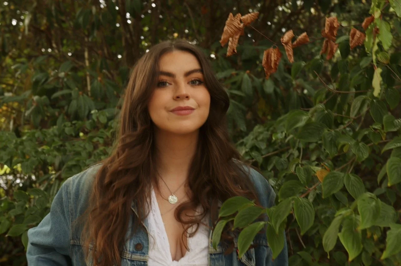 A headshot of Niamh standing outdoors among green foliage. She is wearing a denim jacket, a light shirt, and a silver necklace.