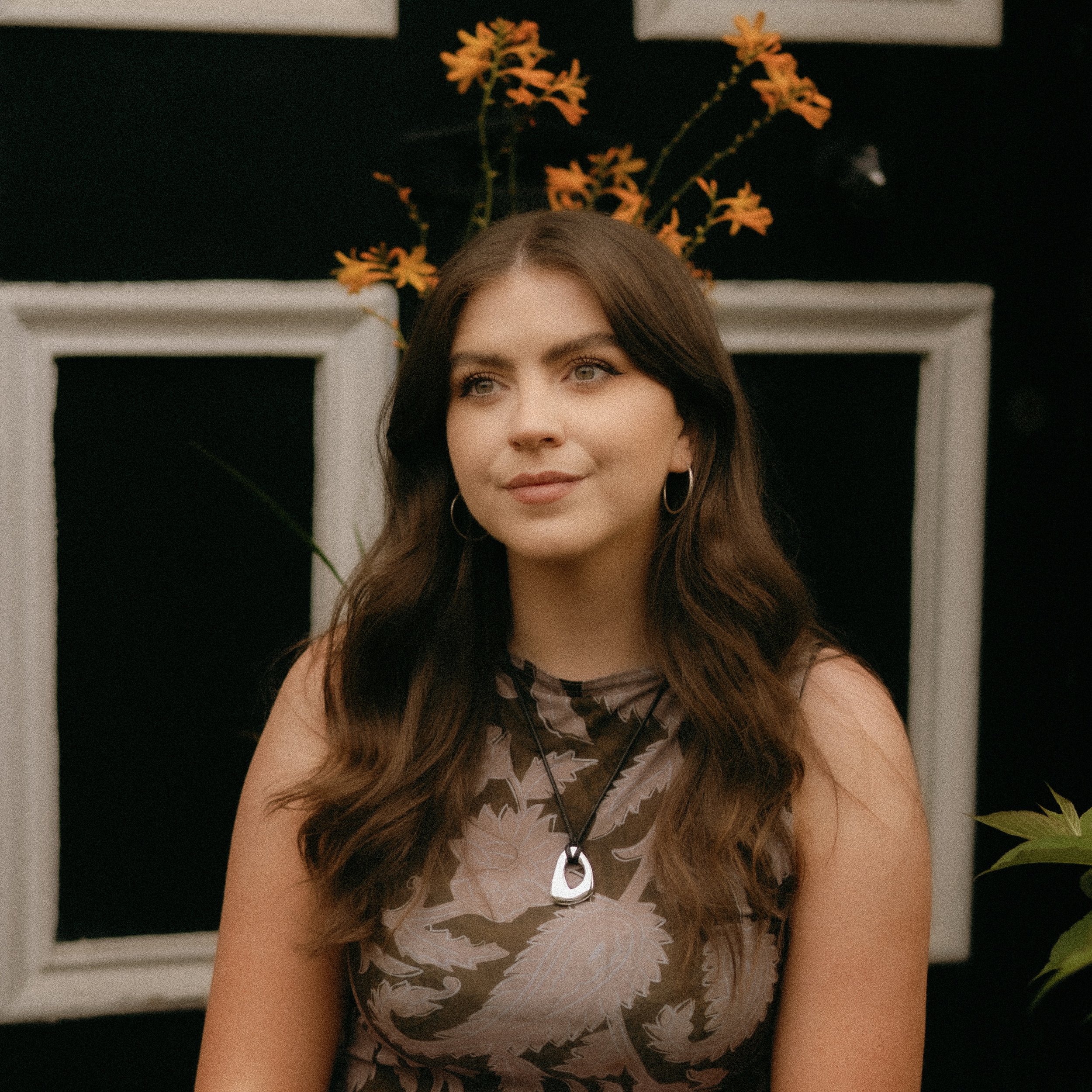 A headshot of Niamh wearing a sleeveless floral top and hoop earrings, with a backdrop of plants and a dark wall with white window frames.