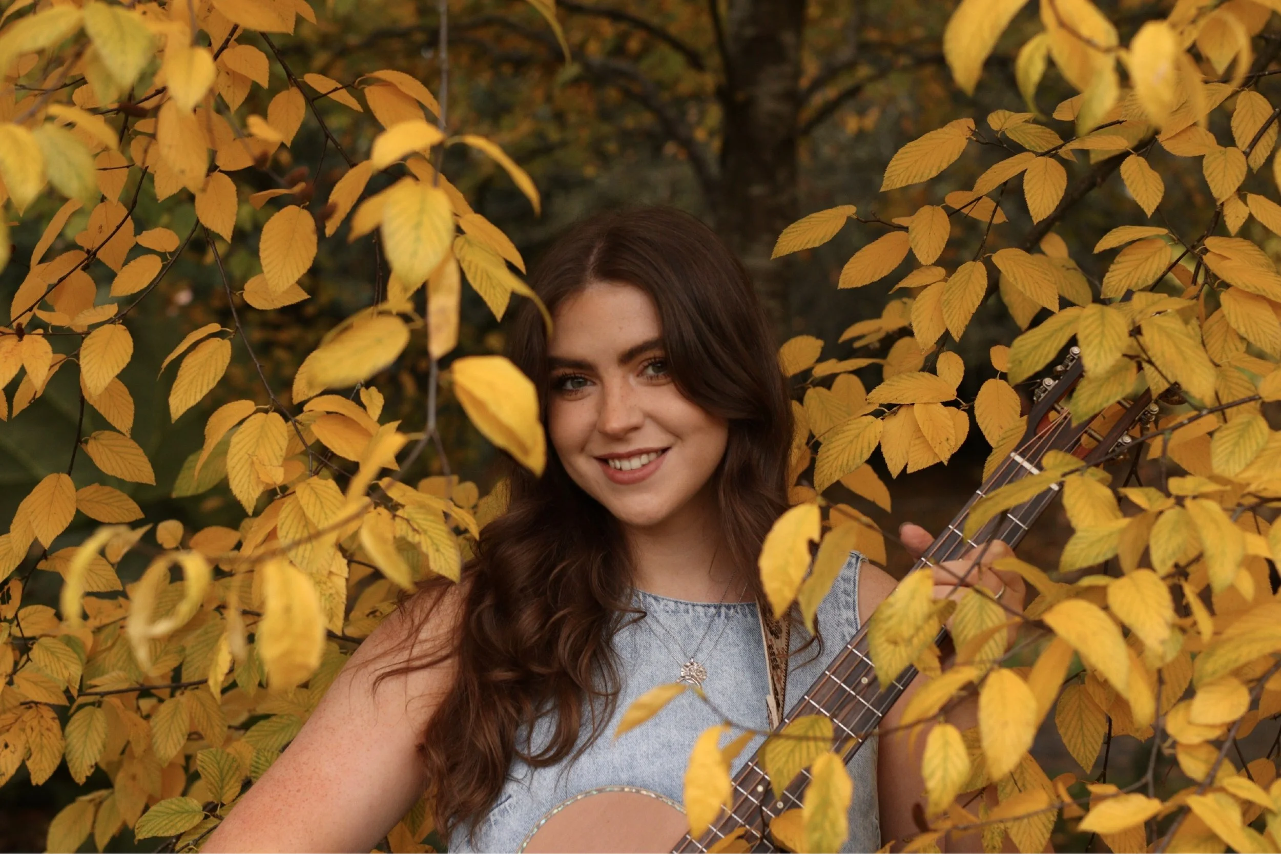 A headshot of Niamh smiling and looking into the camera while holding an acoustic guitar and standing among yellow autumn leaves.