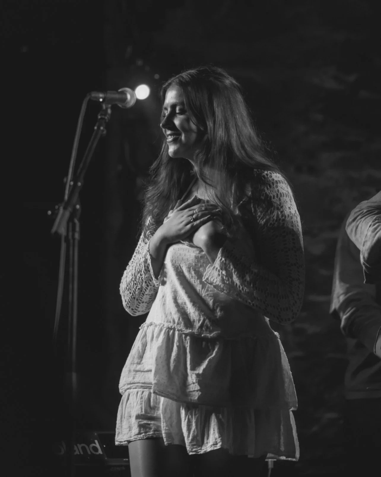 A black and white image of Niamh singing on stage with her hands over her chest and her eyes closed, wearing a light-coloured dress, singing into a microphone in front of her.