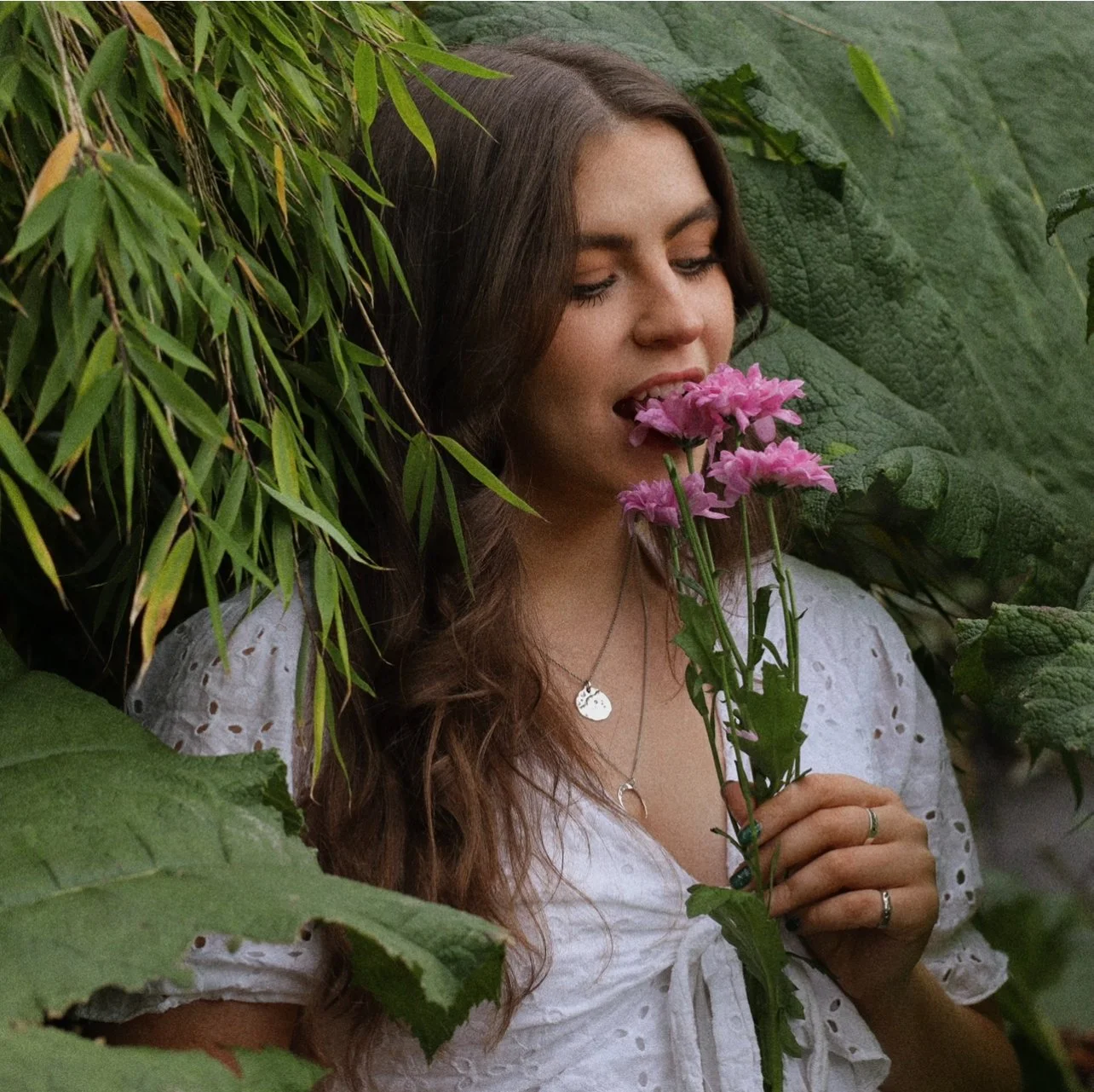 A headshot of Niamh standing among some green leaves and holding a small bunch of pink flowers to her face. She is wearing a light shirt and a silver necklace.