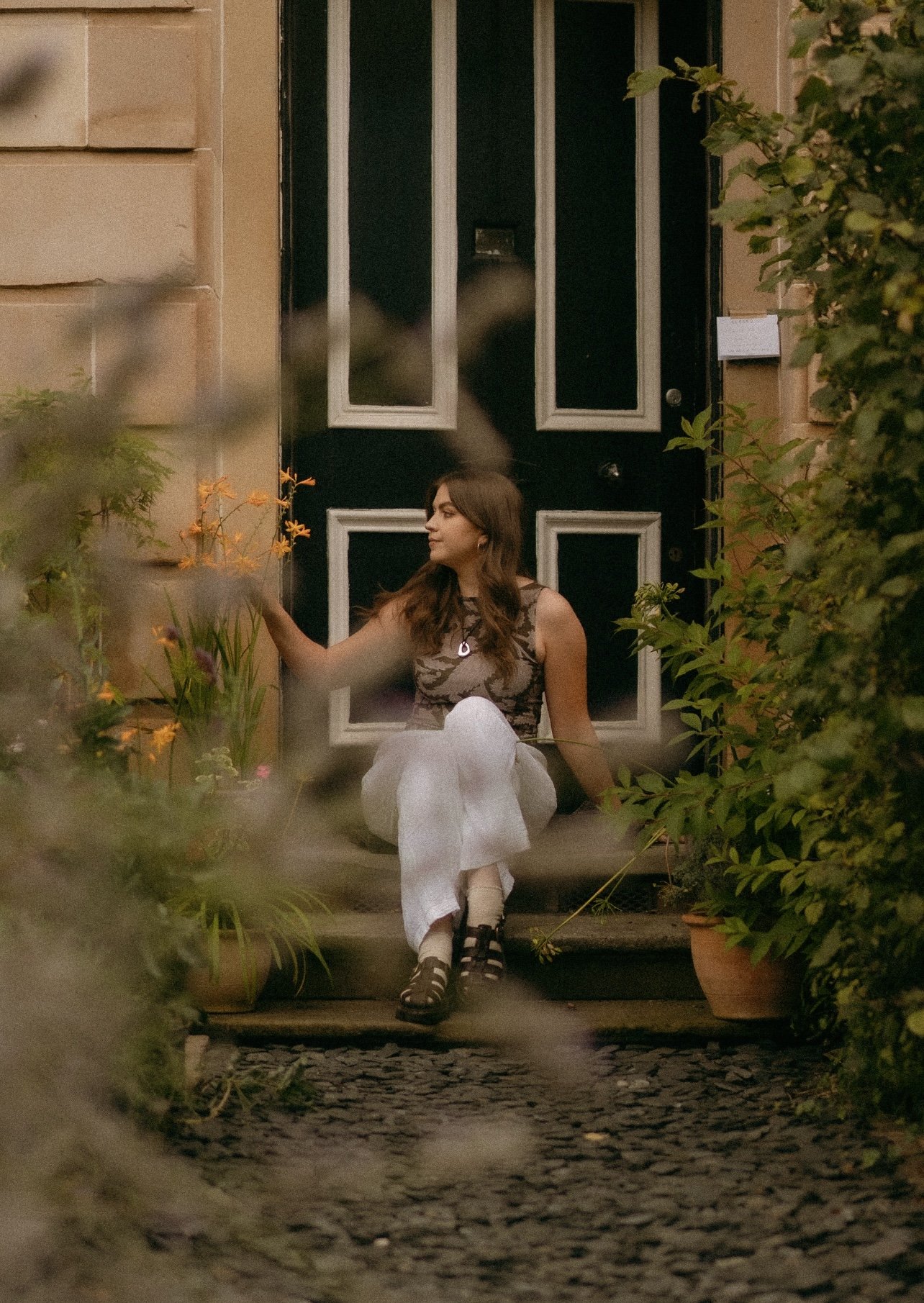 Niamh sitting on some doorsteps outside a house, reaching for nearby flowers, surrounded by plants and greenery. The building has light bricks and a black door with white trimming.