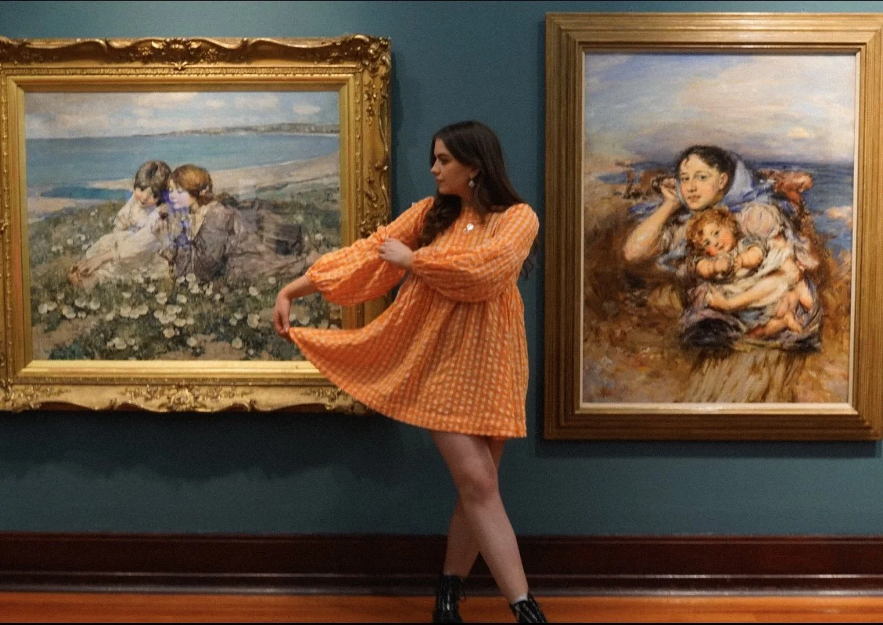 Niamh in an art gallery wearing an orange gingham knee-length dress. She is standing between two framed impressionist paintings, one showing two children sitting on grass and the other showing a boy holding a girl.