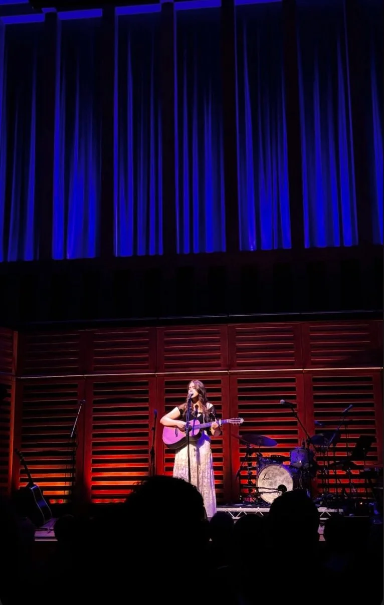 Niamh playing an acoustic guitar and singing on stage with red and purple lighting, a drum set, and other musical instruments behind her, in front of an audience.