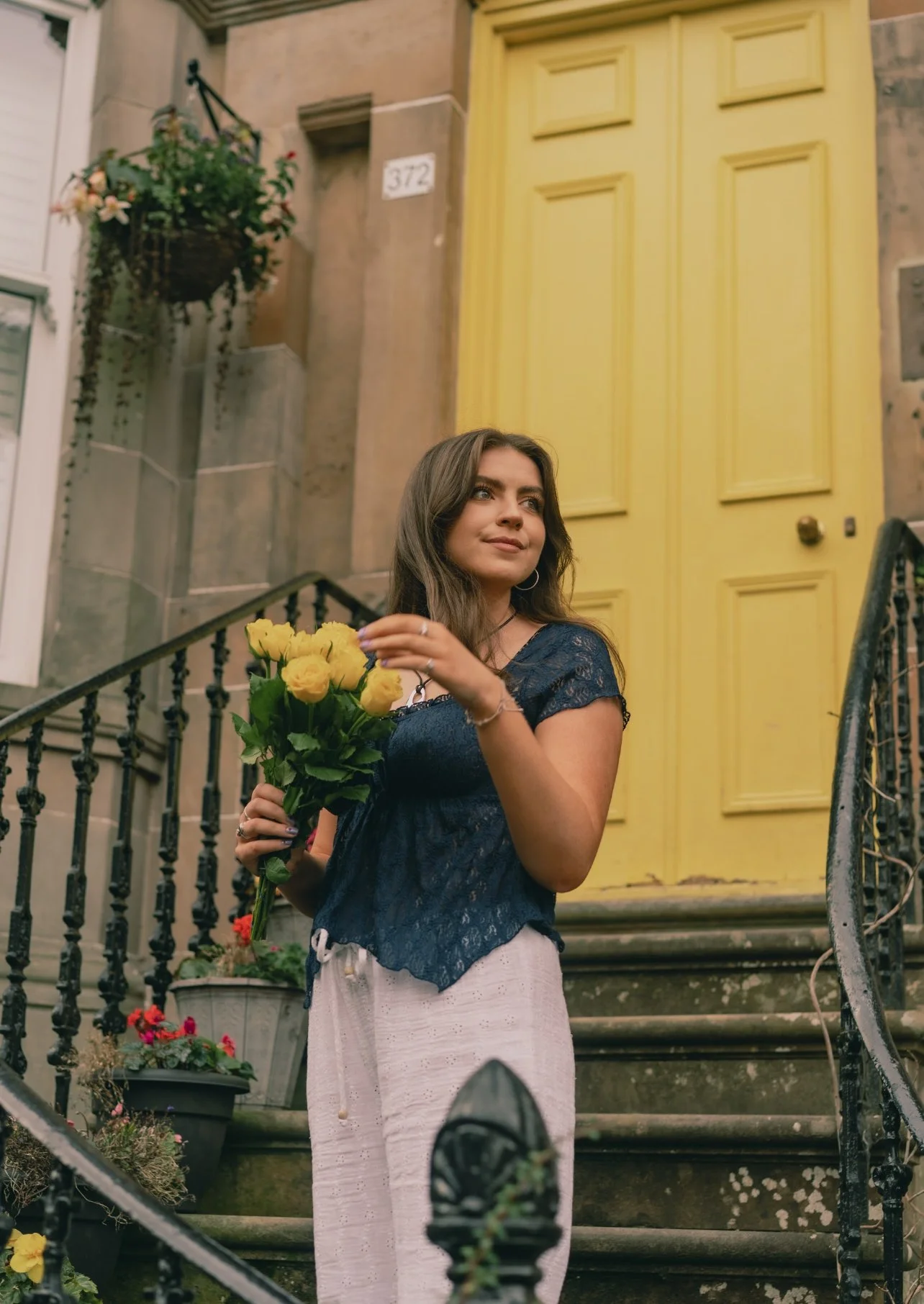 Niamh standing on stairs holding a bouquet of yellow flowers in front of a yellow door with house number 372.