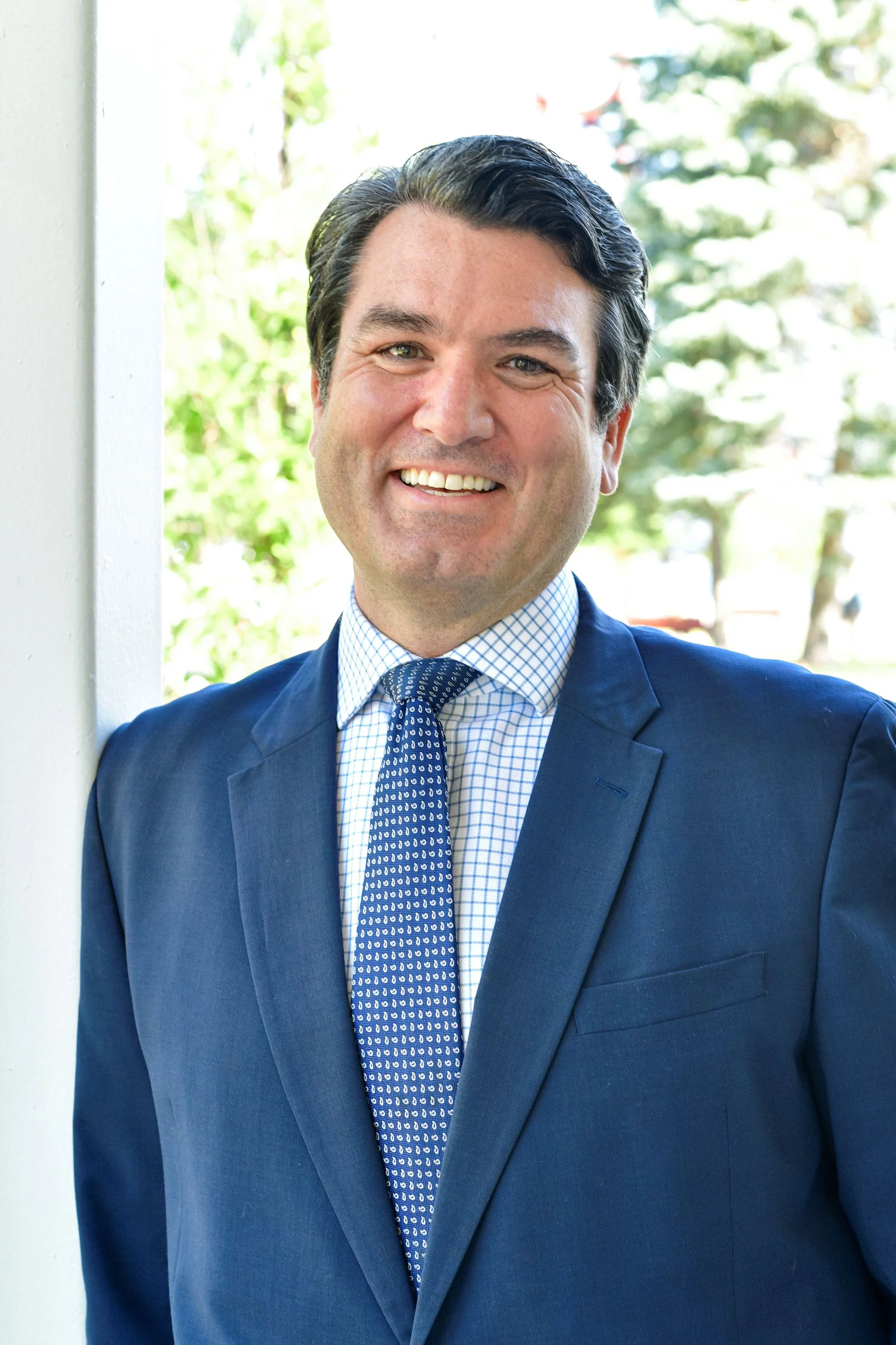 A smiling man in a blue suit with a checkered shirt and patterned tie, standing outdoors near a white wall with trees blurred in the background.