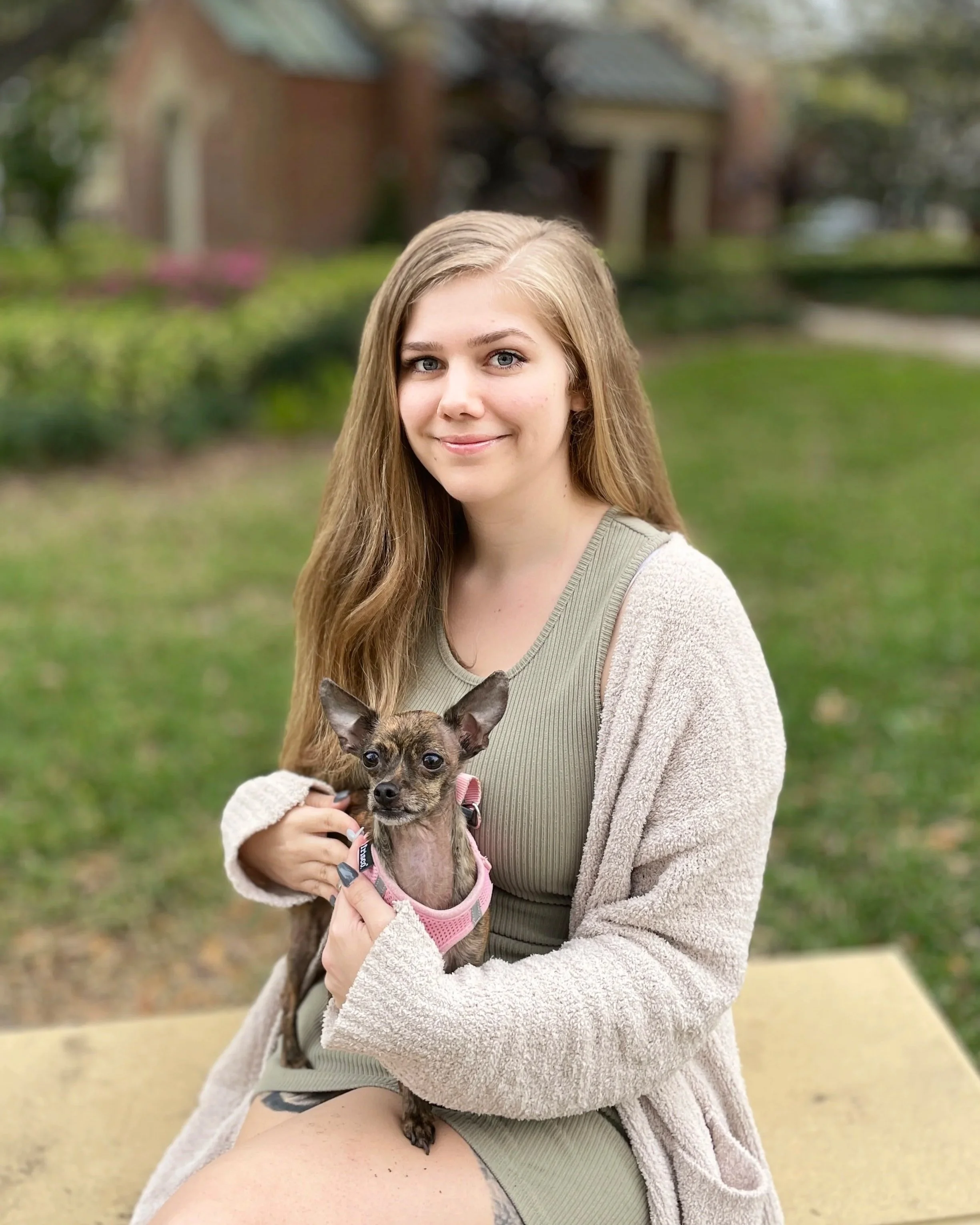 Young woman with long blonde hair smiling and holding a small dog in a park.